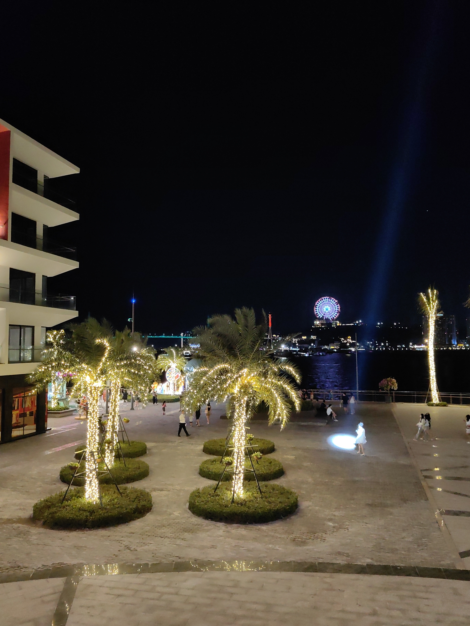 The image depicts a nighttime scene in Bai Chay Ward, Vietnam. In the foreground, illuminated palm trees stand on manicured grounds, bordering a modern building on the left. People are strolling and congregating in the well-lit pedestrian area. In the distance, across a body of water, a Ferris wheel and other structures are visible, also illuminated. A bright beam of light shines vertically into the sky. The overall scene conveys a vibrant, urban waterfront atmosphere.