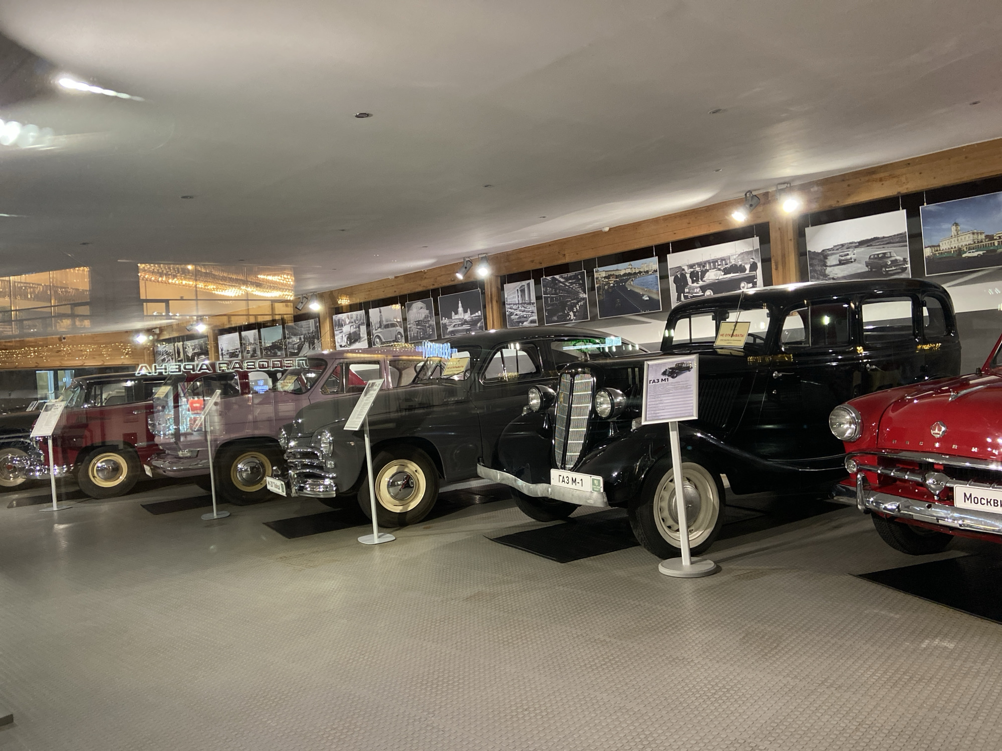 The image shows a collection of vintage automobiles displayed in what appears to be a museum or exhibition hall. The cars are lined up in a row, with information plaques placed next to each one. The hall has a modern interior with a light-colored floor and a ceiling with recessed lighting. A series of black and white photographs adorn the upper portion of the wall, adding a historical context to the vehicle display. Based on the signs accompanying the vehicles, it is possible this is taken in the city of Peresvet, Russia.