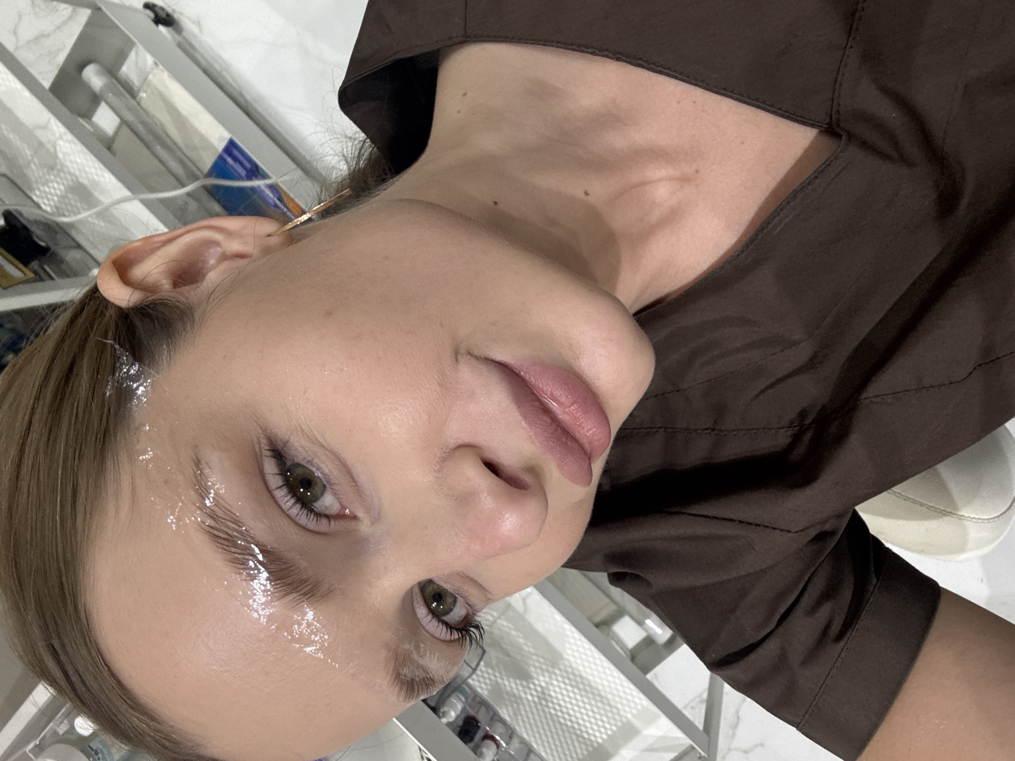 A close-up, slightly angled, eye-level shot shows a woman's face. Her face is pale and shiny, suggesting the presence of a skincare product or moisturizer. Her lips are light pink. She has green eyes and light brown hair, a strand of which is held back by a gold-colored hair pin near her ear. She is wearing a brown collared top. The background is somewhat blurred, and contains medical looking equipment and furniture. The specific location within Budva, Montenegro, is not discernible from this image.