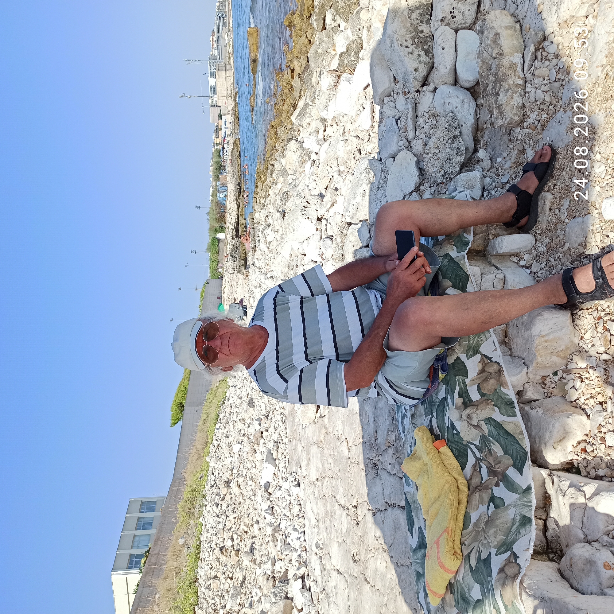A man, appearing to be of Caucasian descent, with white hair and wearing sunglasses, a light-colored baseball cap, a horizontally striped t-shirt, and light-colored shorts, is seated on a blanket laid out on rocks. He is holding a black smartphone in his right hand. The scene takes place on a rocky shore with the blue sea visible in the background. In the distance, buildings and what appear to be people swimming or relaxing near the water can be observed. A yellow towel is folded on the blanket next to the man. The timestamp "24.08.2026 09:53" is overlaid on the bottom right of the image. The general environment suggests a coastal location, likely in the city of Molfetta, Italy, given the architectural style of the distant buildings which are characteristic of the Molfetta promenade.