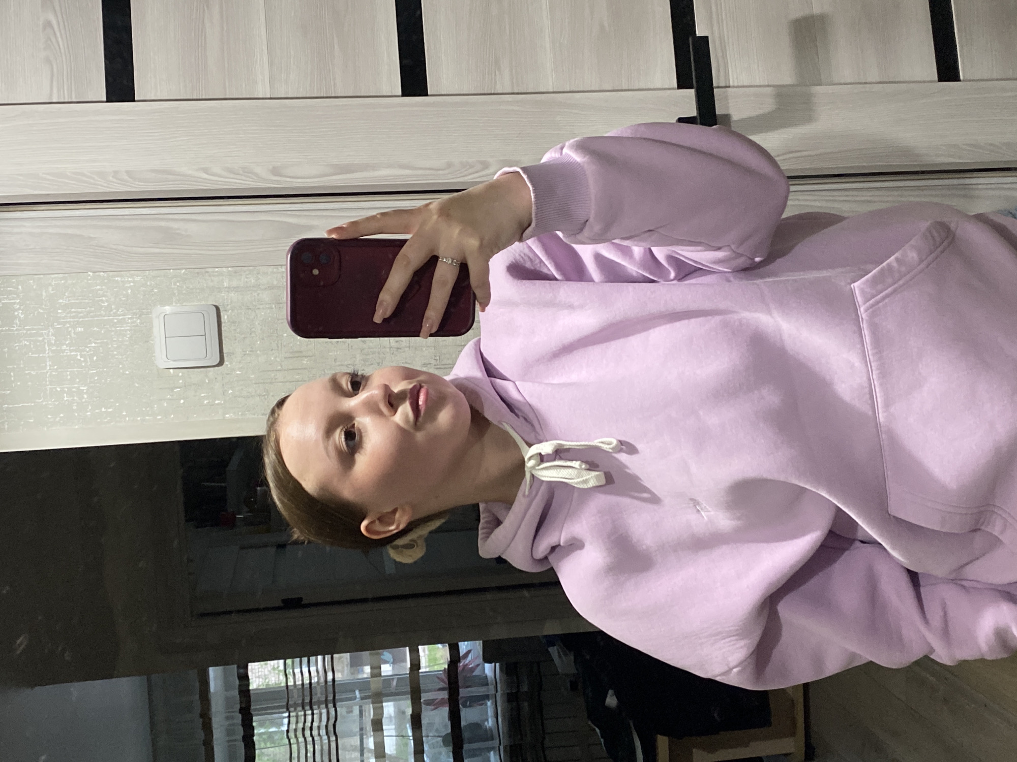 A young woman is taking a selfie in what appears to be an apartment. She is holding a maroon-colored smartphone in her right hand. She has fair skin and her hair is pulled back. She is wearing a light purple hoodie. Behind her is a window with thin, vertically striped curtains, and an out-of-focus view through the window showing trees. Part of a door with vertical dark stripes is visible above her head. A light switch is visible on the wall. Based on visual evidence, the location is inconclusive as to being in the city of Chaikovsky, Russia.