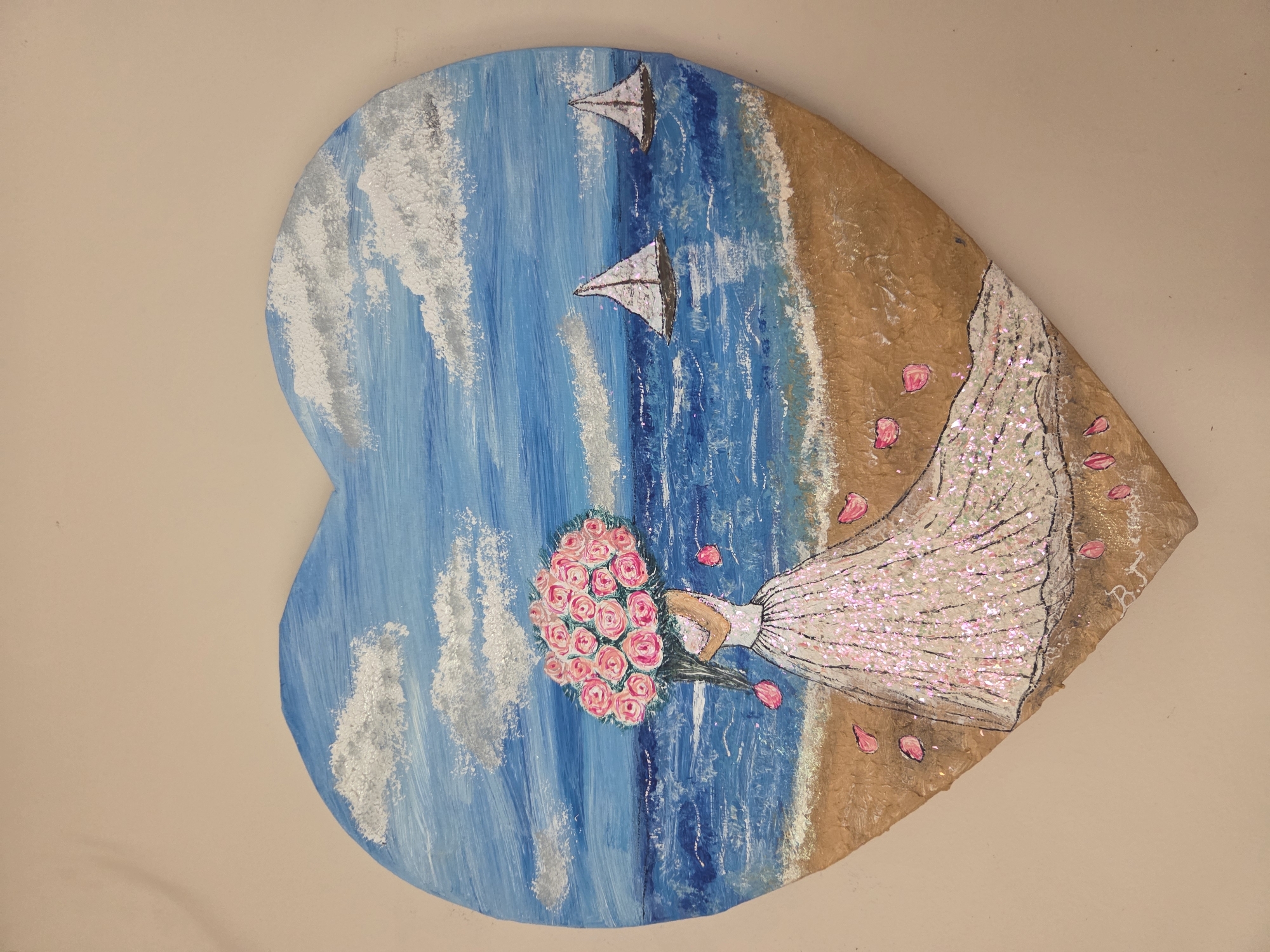 The image shows a heart-shaped painting or artwork hanging on a beige or light tan wall. The artwork depicts a beach scene with a bride. The left side of the heart shows a blue sea with white, foamy waves and two sailboats in the distance. The right side shows a tan-colored sandy beach. A woman, presumably a bride, is depicted in a shimmering white gown, holding a bouquet of pink roses. The gown appears to have glitter or sequins. A few loose pink petals are scattered near the bride and the beach. A signature, "B.J," is visible on the right edge of the heart near the dress. The overall scene evokes a wedding theme with a beach backdrop. The location is not evident in this image.