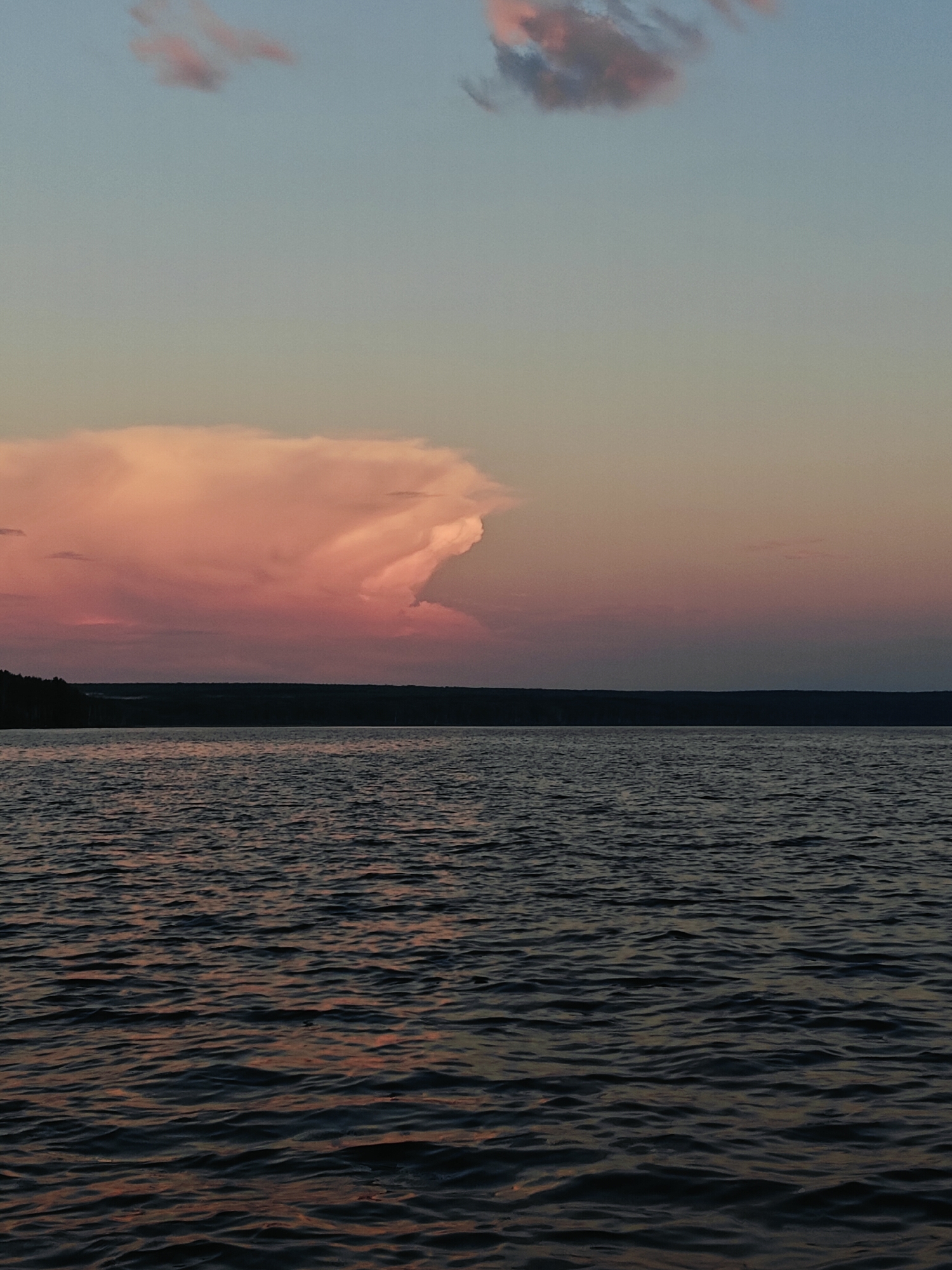 The image depicts a serene sunset over the Kama River in Chaykovsky, Russia. The sky transitions from a soft blue at the top to hues of pink and orange near the horizon, illuminated by a large, fluffy cloud formation. A dark, silhouetted treeline borders the far bank of the river. The water's surface is calm, with gentle ripples reflecting the subdued colors of the sky. There are no people or man-made structures visible in the frame, emphasizing the natural beauty of the landscape.