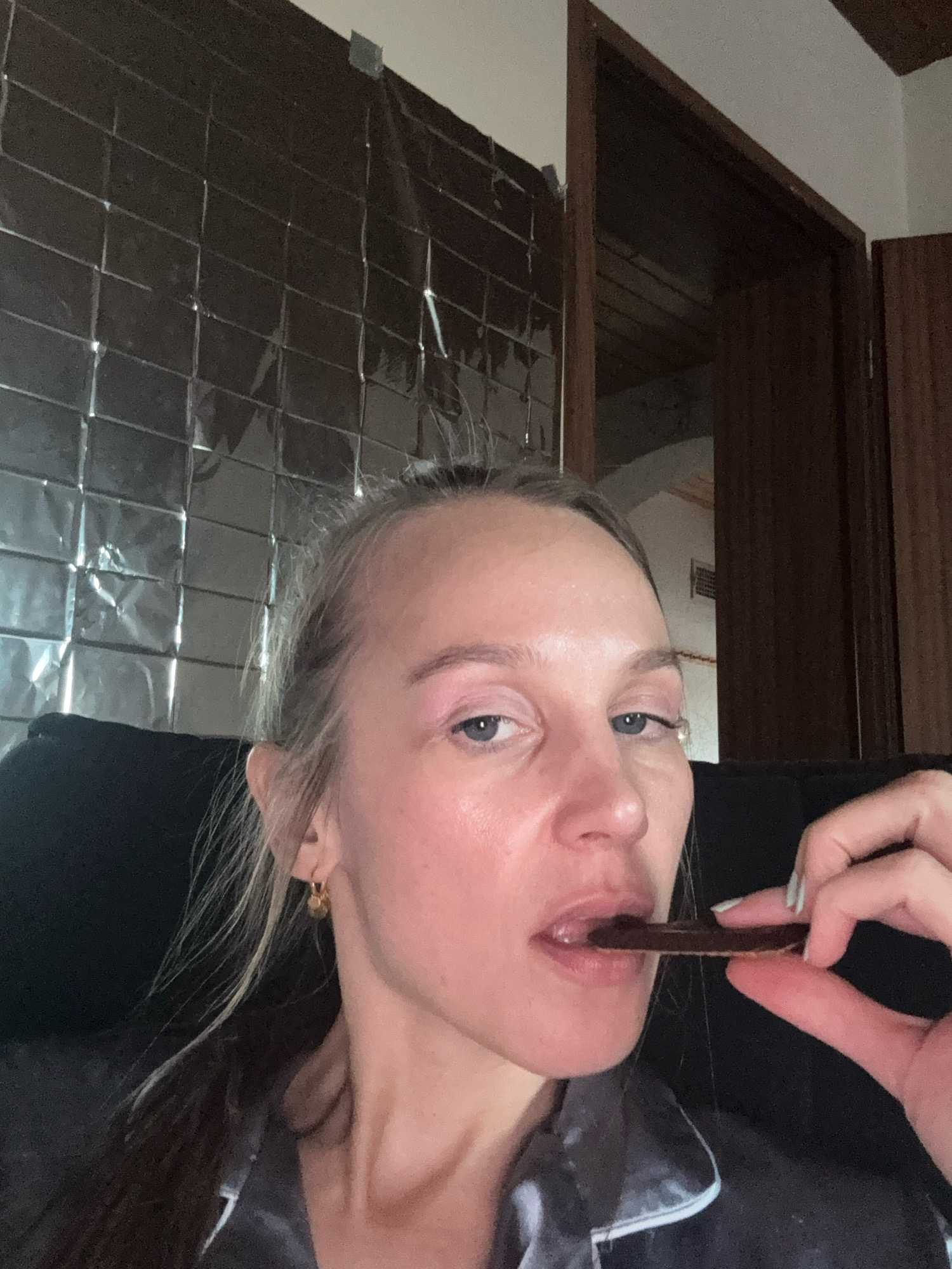 A woman is pictured close-up, eating a thin, dark chocolate cookie. Her hair is pulled back, and she is wearing a grey pajama top. In the background, a wall covered in reflective squares and a wooden doorway are visible. The scene appears to be indoors, likely in a residential setting in Überherrn, Germany.