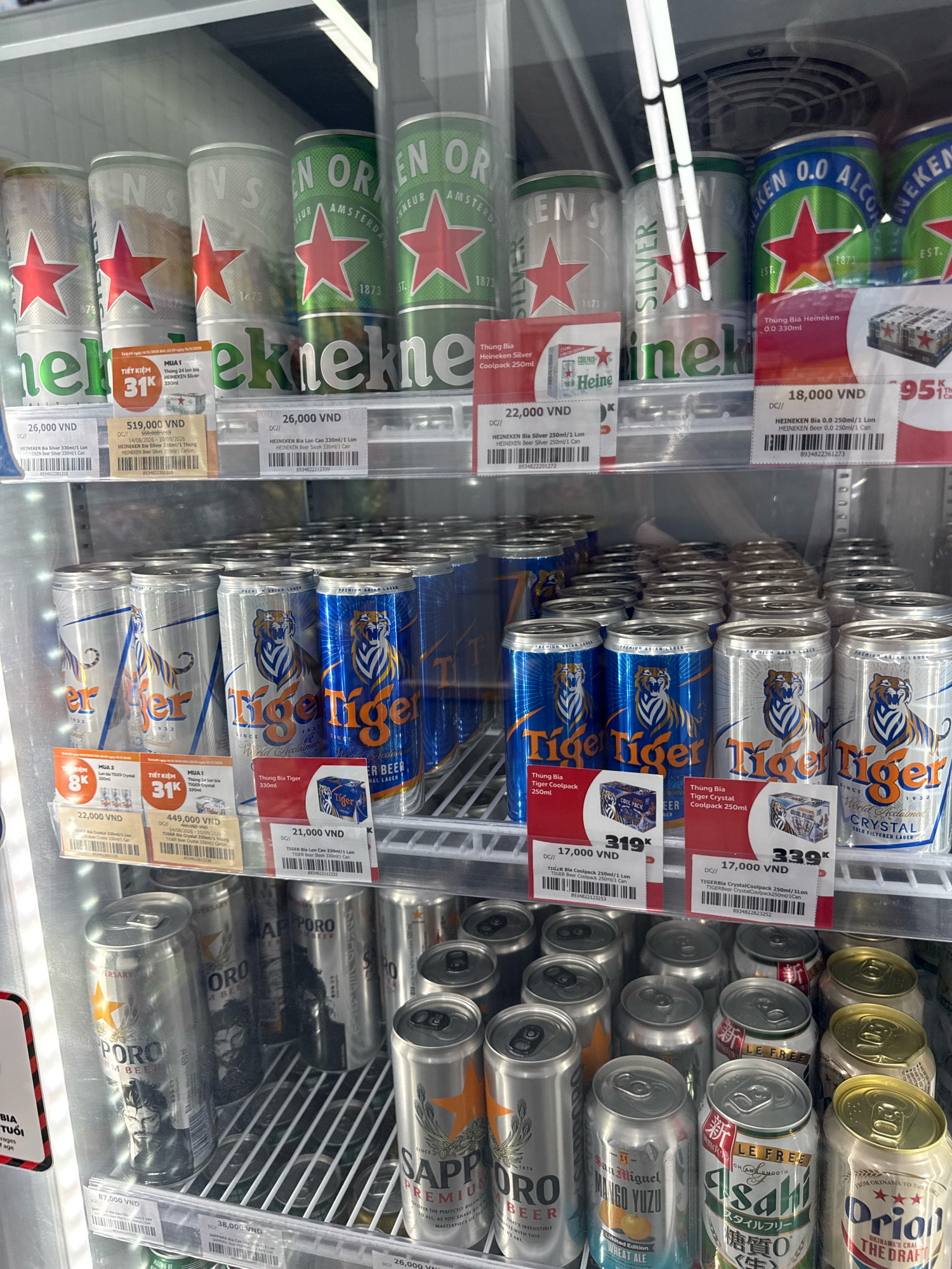 The image is a close-up, eye-level, interior shot of a refrigerated display case in a convenience store in Nha Trang, Vietnam. The case is stocked with a variety of canned beverages, predominantly beer. In the upper shelves, several cans of Heineken are visible, including Heineken Silver and Heineken 0.0. There are also cans of Tiger beer, both regular and crystal. The lower shelves are filled with more Tiger beer cans, as well as cans of Sapporo Premium Beer, San Miguel Mango Yuzu Wheat Ale, and Orion The Draft. Price tags are visible for many of the products, indicating prices in Vietnamese Dong (VND). The lighting is artificial and bright, reflecting off the metal cans and the glass of the display case. No people are visible in the image. The overall impression is of a well-stocked beverage cooler in a commercial setting.