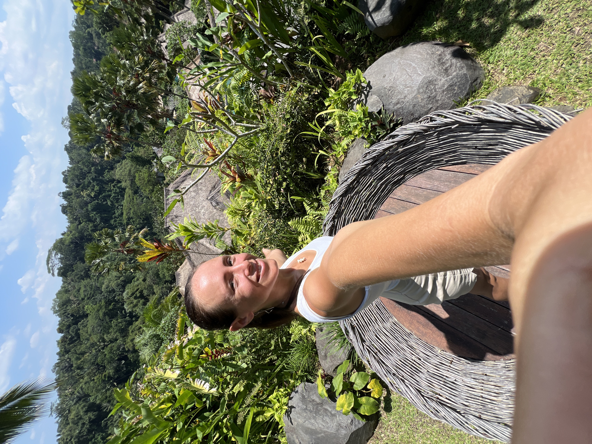 A woman is taking a selfie outdoors in Kedewatan, Indonesia. She is wearing a white tank top and shorts, sitting in a large, circular chair with a wooden base and a woven wire-like back. The foreground includes lush green foliage, with various plants and trees surrounding the chair. Several large, dark grey rocks are scattered around the seating area. In the background, there's a dense, green forest covering the hillside. Above the forest, there's a blue sky with scattered white clouds. The location is identifiable as being within the Ubud area of Bali, specifically Kedewatan, due to the distinctive tropical vegetation and hilly landscape. The image appears to be taken during daylight hours.