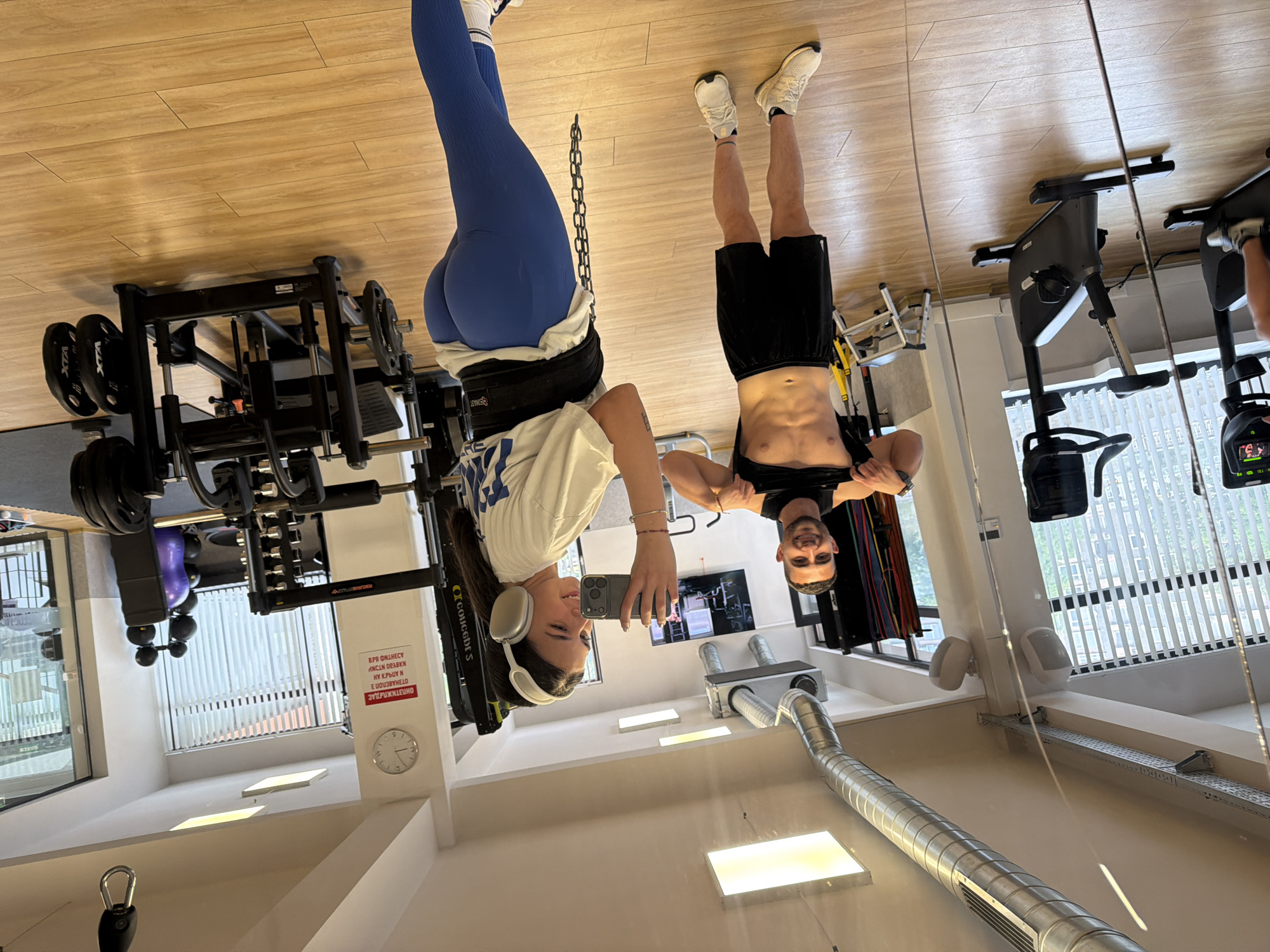 Two people hang upside down in a gym, suspended by their ankles. On the left, a woman in blue leggings, a white t-shirt, and white sneakers is wearing headphones and holding a cell phone. She is attached to the equipment by an ankle harness. On the right, a shirtless man in black shorts and white sneakers is also hanging upside down. They are taking a selfie. The gym equipment includes a weight rack, a rowing machine, and stationary bikes. The gym has wood-look floors, white walls, and large windows. A sign in Cyrillic is visible on the wall, identifying the location as being in Gabrovo, Bulgaria.