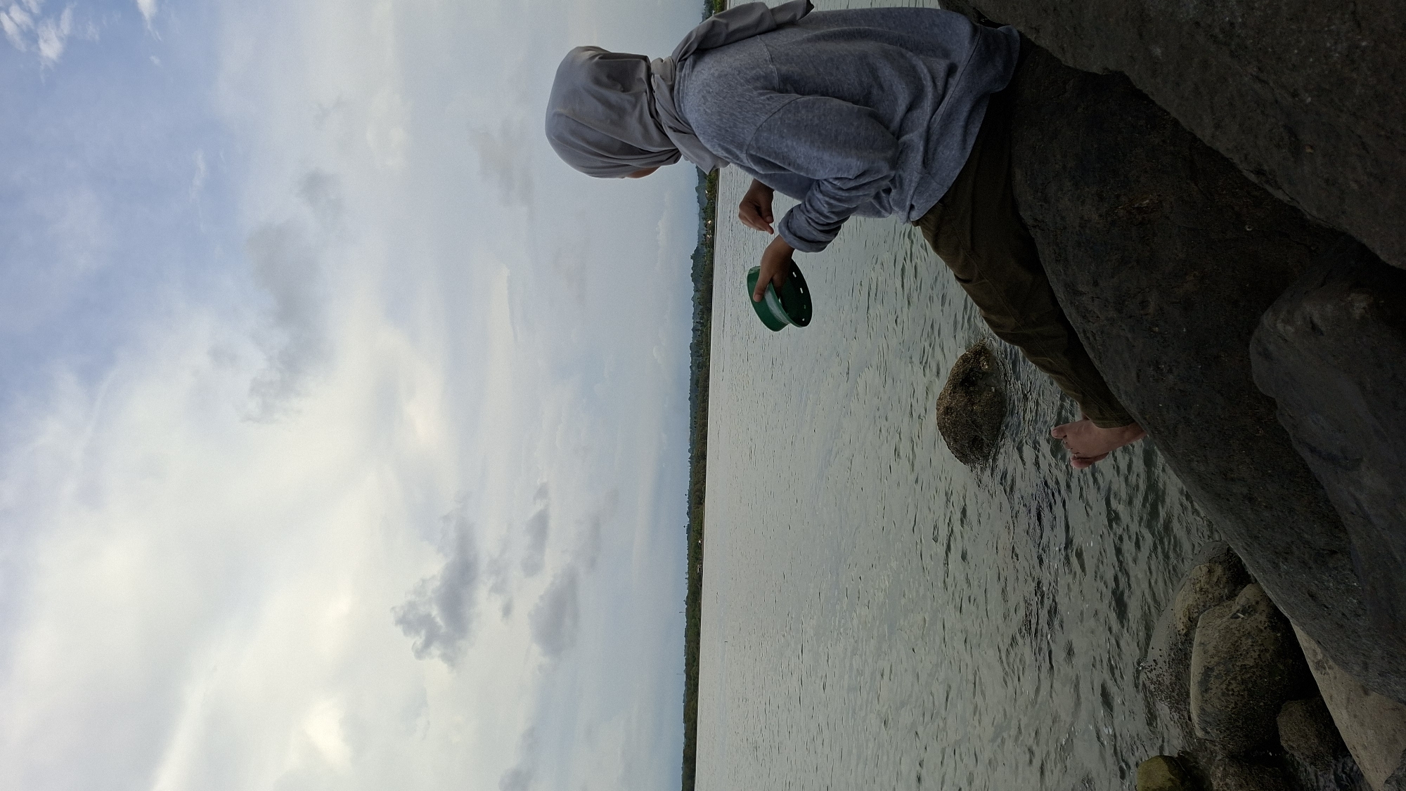 A person wearing a grey hijab and a grey hooded sweatshirt sits on large rocks by the water, holding a green object. The person is facing away from the camera, looking out at the water and a distant treeline under a cloudy sky. The water is calm, with ripples on the surface. Rocks are visible in the foreground and extending into the water. The location appears to be a coastal area, possibly a bay or estuary, in Lhokseumawe, Indonesia. The atmosphere is subdued, suggesting overcast weather.