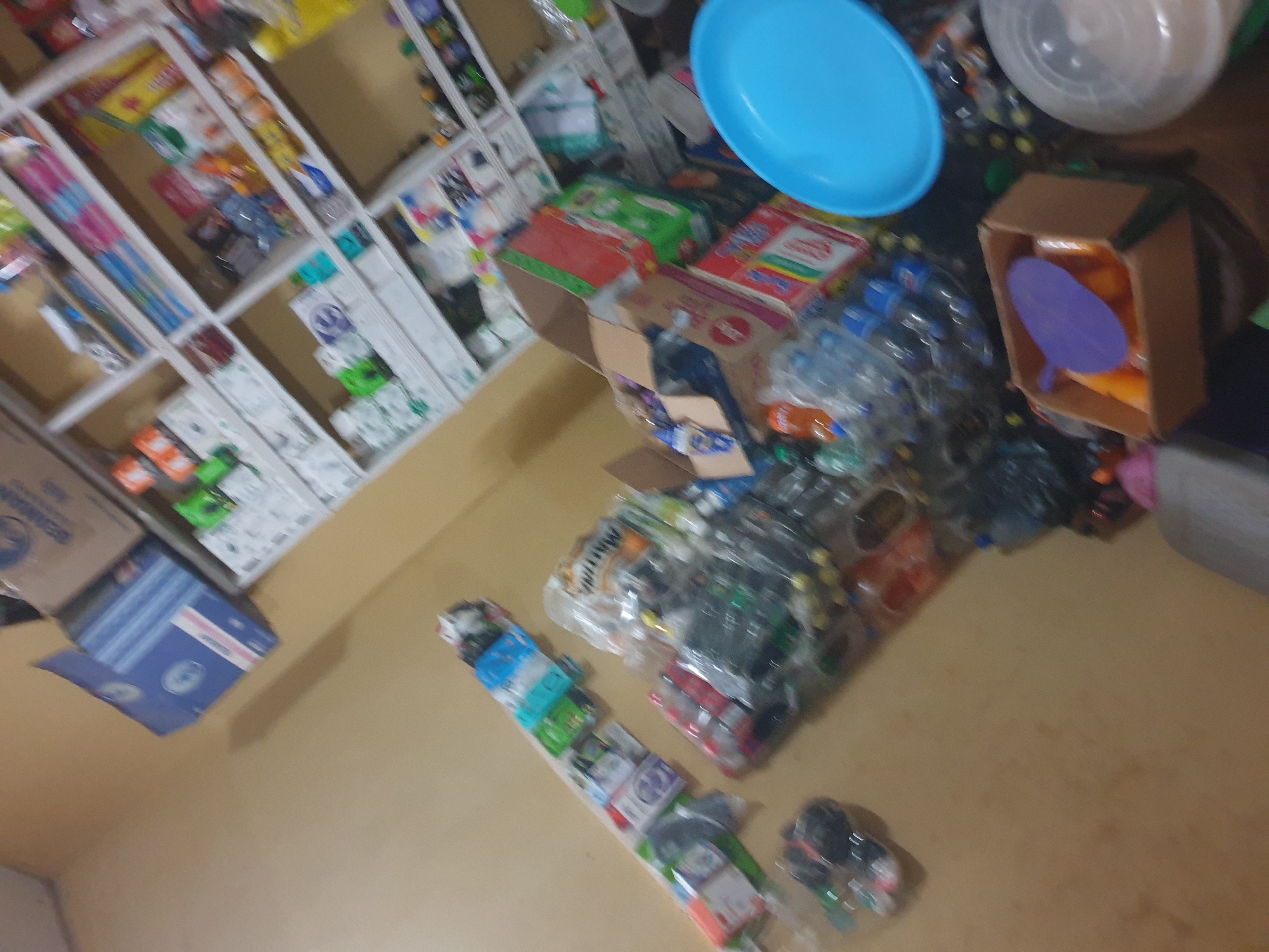 This is a photograph of a cluttered room, possibly a storeroom or a small shop, located in Anilelerin, Nigeria. The room is filled with various items, including shelves stocked with packaged goods, boxes, and plastic bottles. A wall is visible on the left side of the image, painted in a light yellowish-brown hue. On the floor, there are stacks of plastic bottles and boxes. In the foreground, a light blue plastic plate or tray is prominently displayed. The overall impression is one of disarray, with items piled up and shelves overflowing. The lighting suggests it might be indoors, possibly with artificial lighting. No people are visible in the photograph.