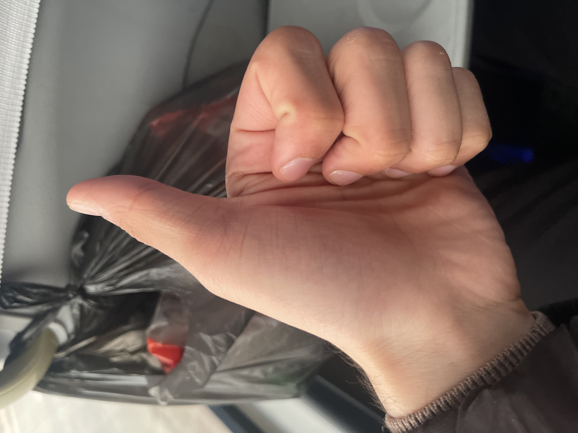 A close-up photograph depicts a person's left hand, with the fingers curled into a fist except for the extended thumb, positioned against a blurred background. The hand is pale, showing skin texture and nail detail. The wrist is partially visible, revealing a brown knitted sweater cuff. Behind the hand is a black plastic bag containing indiscernible items with a glimpse of red. The background also includes a white surface, potentially a car interior element. The setting is not identifiable as being in Karmana, Uzbekistan, based on the provided image.