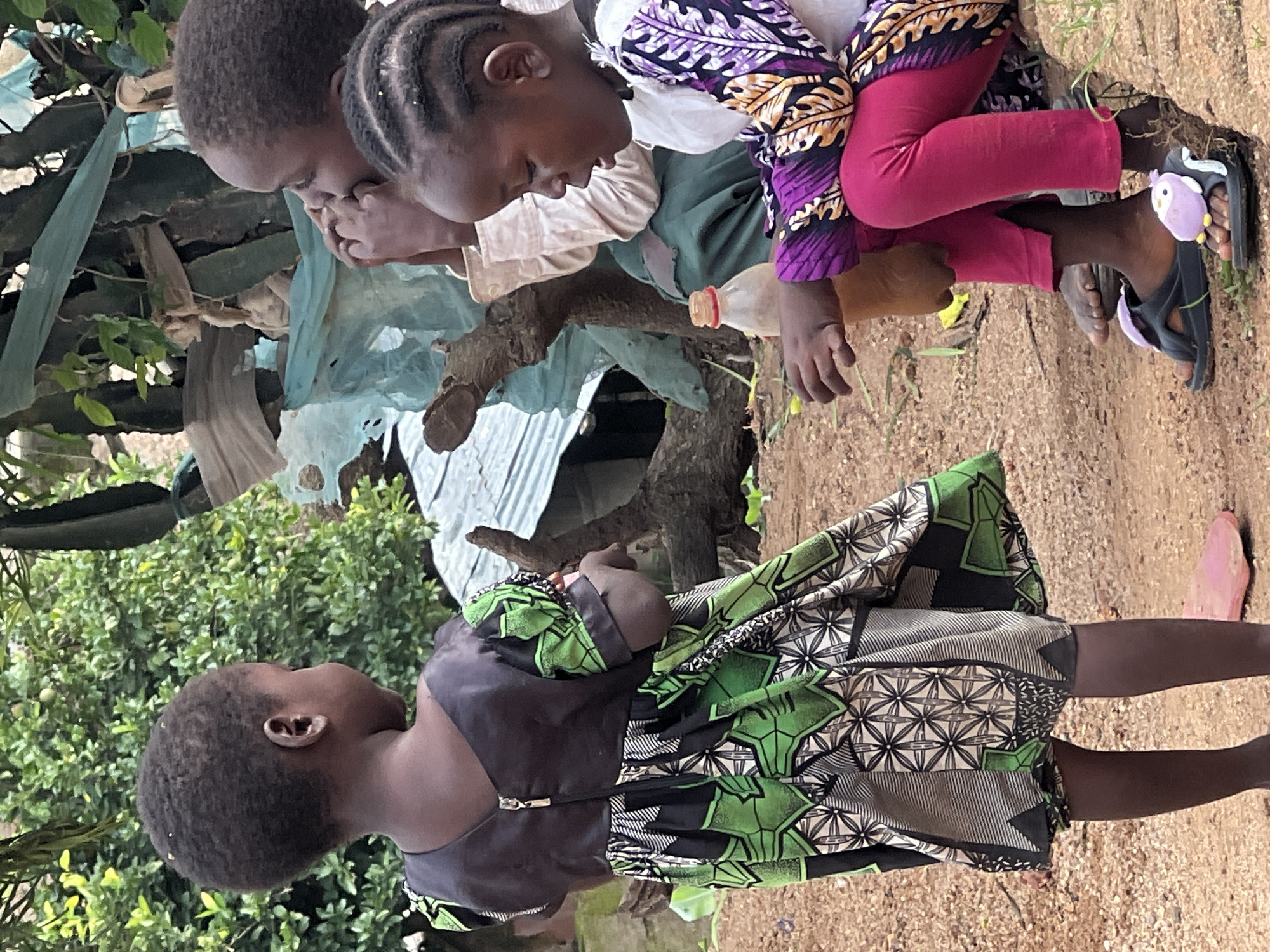 In Jos, Nigeria, three young girls are gathered outdoors on a dirt ground. The girl on the left, facing away from the camera, wears a black top with a green and black patterned dress featuring geometric designs and a zipper closure. The girl in the middle, partially visible, is positioned behind the other two and is seen touching her face. The girl on the right, facing left, is wearing a white top with a brightly colored, patterned sleeve and pink leggings. She is wearing black sandals adorned with purple owl-shaped charms. A red-capped bottle, possibly containing liquid, rests on the ground near her. The background consists of lush green foliage and a makeshift structure with a corrugated metal roof, suggesting a rural or semi-urban setting. The overall atmosphere appears casual and communal.