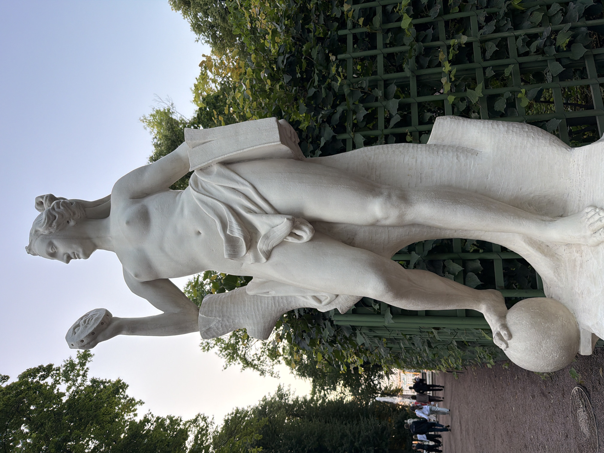 A full-body white marble statue of a nude figure stands in a park. The figure is male, with long hair styled in curls, and is posed with its left foot resting on a sphere. The figure holds a circular object with a relief face in its raised left hand and a rectangular object, possibly a book, in its right hand. The statue is situated outdoors, with a gravel pathway, people walking, green foliage, and a green lattice fence in the background. The sky is visible above. The statue is located in the Summer Garden in Saint Petersburg, Russia.