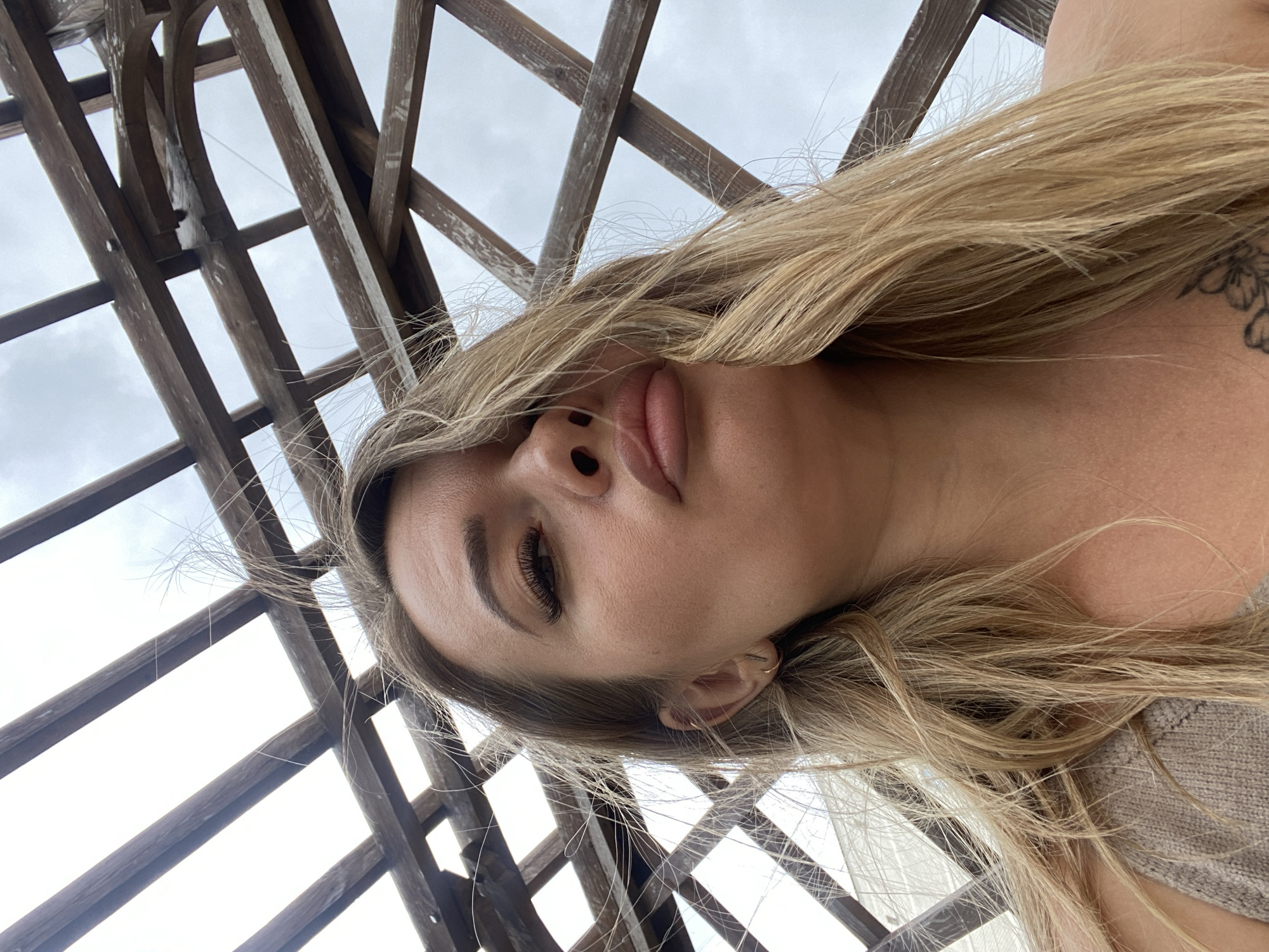 A woman is positioned under a wooden pergola-like structure, which is visible against a cloudy sky. The woman has long, blonde hair and light skin, and a tattoo is visible on her right shoulder. The photograph appears to be taken from a low angle, looking up at her. It's not possible to determine the location of the image in Izhevsk, Russia.