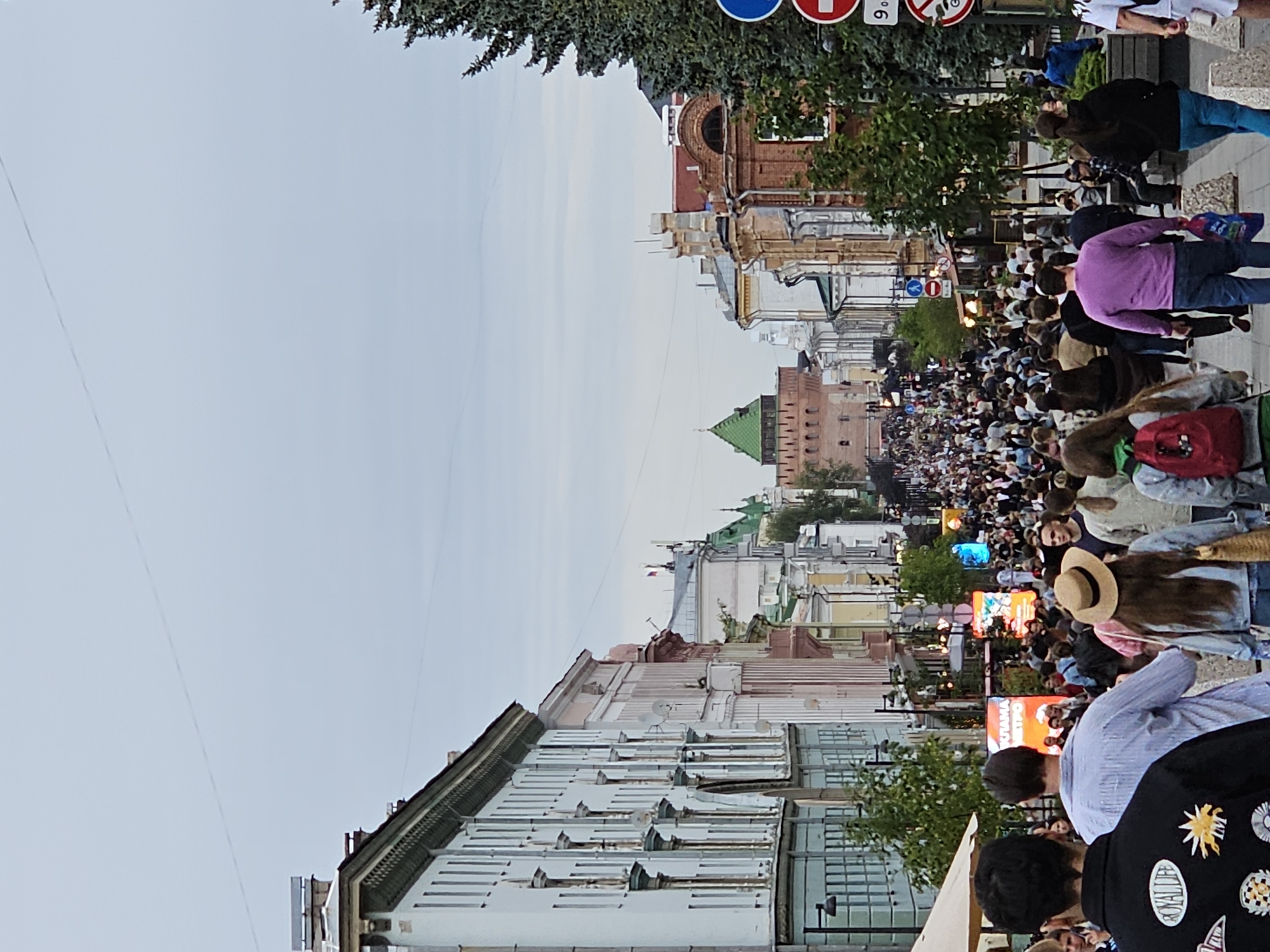 This media file depicts a street scene crowded with people, suggesting a public event or festival. The perspective is from a slightly elevated position, looking down a street lined with buildings on both sides. The buildings appear to be a mix of architectural styles, with some exhibiting older, ornate features and others appearing more modern. Trees are interspersed along the street, and some of the buildings have balconies and windows that suggest residential or commercial use.
The people in the image are diverse, with a variety of clothing and ages. Many are walking, and some appear to be looking at something ahead or interacting with each other. The density of the crowd implies a significant gathering. There are several illuminated signs and displays visible in the crowd, some of which appear to be electronic billboards, displaying colorful graphics and text. One sign in particular seems to be a bright orange with Russian text. The time of day appears to be late afternoon or early evening, given the soft light and the illumination of the signs. The weather seems to be overcast or cloudy.
The scene is suggestive of a lively urban environment where a public celebration or event is taking place. The presence of what looks like a green-domed building in the distance and the architectural style of some of the structures could indicate a historical district. The overall emotion conveyed is one of bustling activity and communal engagement.