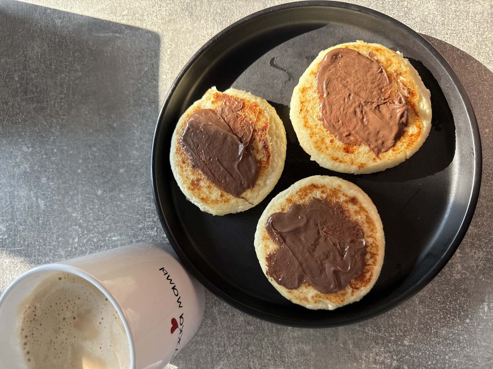 A close-up, overhead shot shows three small, round pancakes topped with chocolate spread on a black plate. The pancakes are golden brown with slightly crispy edges. To the left of the plate, a white mug filled with coffee and a foamy top is visible. The mug has a red heart and black text that appears to read "Пукка мамы". The subjects are placed on a textured, grey surface that resembles a countertop. The lighting suggests it's daytime, with a shadow cast from the right side of the frame. There are no people or animals present. The location is indoors, possibly a kitchen or dining area.