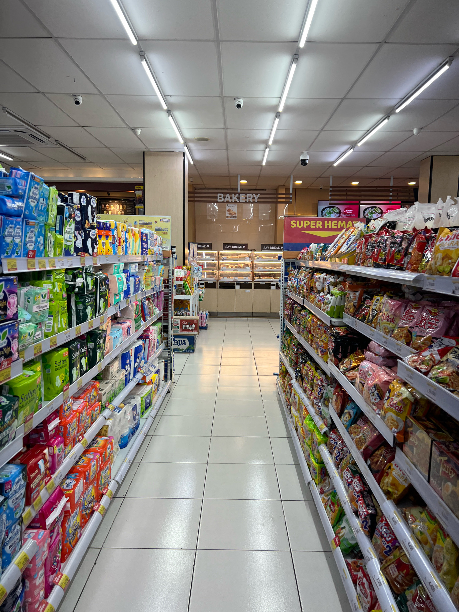 The image depicts the interior of a well-lit convenience store or minimarket, likely a branch of a chain such as Indomaret or Alfamart, given the typical layout and product displays. The perspective is from the aisle, looking forward towards the back of the store.
The store features rows of shelves stocked with various consumer goods. On the left, the shelves are filled with hygiene products, including packaged sanitary pads and diapers, with colorful packaging featuring brand names like "Softex" and "Pampers." Further down the aisle, there are more packaged goods. On the right side, the shelves are laden with snacks, chips, and instant food products, with prominent brands and colorful packaging. Signs on the shelves indicate prices.
In the background, there is a bakery section with display cases filled with baked goods. Above the bakery, a sign reads "BAKERY." Further back, a large digital screen displays what appears to be food imagery. A prominent yellow and red sign on the right aisle reads "SUPER HEMAT," which translates to "Super Savings" in Indonesian, suggesting a promotional display.
The environment is clean and organized, with white tiled flooring. Overhead, fluorescent tube lights illuminate the space. Several security cameras are visible attached to the ceiling, indicating security measures. There are no people visible in the frame. The overall impression is that of a typical retail environment in Indonesia, designed for efficient stocking and customer browsing. The specific location in Denpasar, Indonesia, is inferred from the general characteristics of such stores commonly found in Indonesian cities.