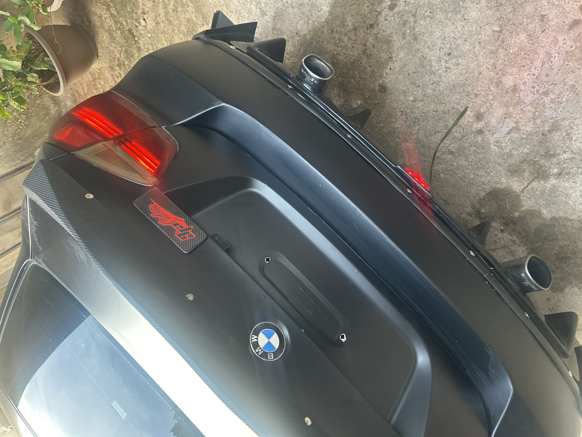 The media file captures a close-up, angled view of the rear of a heavily modified, matte black car, possibly a BMW given the visible emblem. The setting appears to be outdoors on a concrete surface, likely in Barden Yana, Nigeria, with a potted plant visible in the upper left corner.
The main subject is the car's rear, featuring aftermarket customizations. The car has a matte black finish on its bumper and what appears to be a rear diffuser with prominent fins and two circular exhaust tips. One exhaust tip clearly shows the brand name "BORLA" and "PERFORMANCE" inscribed on it. The taillights, visible on the left side of the image, are illuminated with distinctive red LED strips. The trunk lid section features a carbon fiber-look wrap and a circular BMW emblem with the letters "BMW" around it. To the left of the emblem, a red and black sticker is affixed, displaying a stylized logo and the text "ACUITY PERFORMANCE". The rear window is partially visible, reflecting the sky and some white elements, likely a stripe on the car itself.
No human activity is taking place; the car is stationary. The lighting suggests it is daytime with clear weather. The overall impression is of a customized performance vehicle parked in a casual outdoor setting.