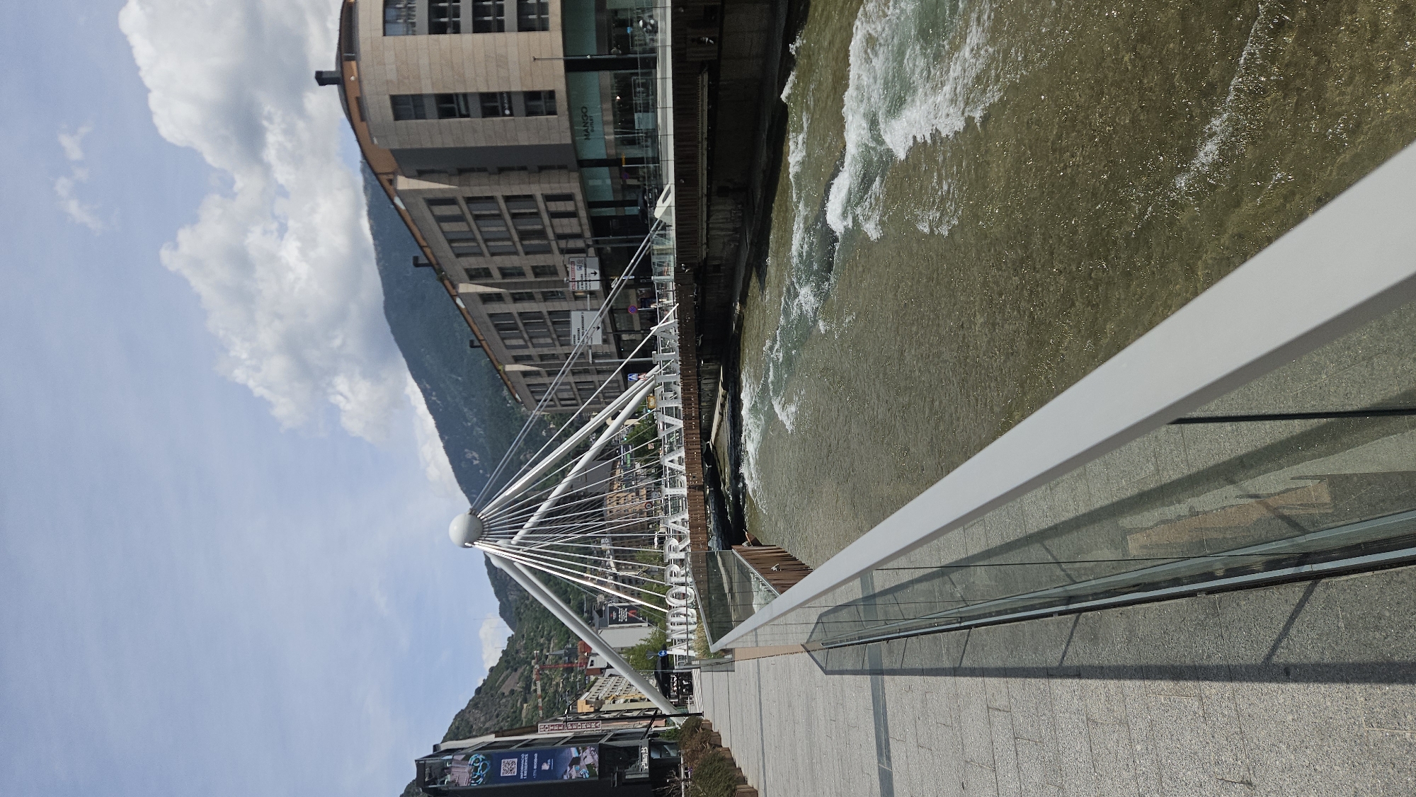 The image captures a detailed perspective of a city scene in Andorra la Vella, Andorra, under a bright, partly cloudy sky. The vantage point is from a paved stone walkway along the left bank of a river, looking towards a distinctive pedestrian bridge and the urban landscape beyond.
In the immediate foreground on the left, a light grey handrail with clear glass panels separates the granite-tiled walkway from the river. Shadows from the railing and an unseen object are cast on the walkway. The river, likely the Gran Valira, flows from the upper left towards the lower right, displaying visible rapids and white water indicating a moderate current; its water is a murky greenish-brown.
The central feature is a modern pedestrian bridge spanning the river. It is characterized by a prominent white spherical pylon in its center, from which numerous white suspension cables radiate downwards to support the brown-colored bridge deck. Large, white, capitalized letters spelling "ANDORRA LA VELLA" are affixed to the side of the bridge structure, facing the viewer. Several pedestrians are visible walking across the bridge.
Beyond the bridge on the left, multi-story commercial buildings with modern facades line the street, displaying storefronts and promotional banners. Trees and vegetation are visible on the lower slopes of the mountains behind these structures. On the right side of the river, across from the walkway, a row of contemporary multi-story buildings with large windows and light-colored facades (beige/tan) are visible. One building at a corner features a rounded architectural element. A prominent "MANGO OUTLET" sign is visible on a ground-level storefront, and various road signs are present further down the street.
The background is dominated by steep, tree-covered mountains that rise directly behind the city structures. The sky above is a light blue with scattered white, cumulus clouds. The scene is illuminated by strong daylight, creating clear shadows.