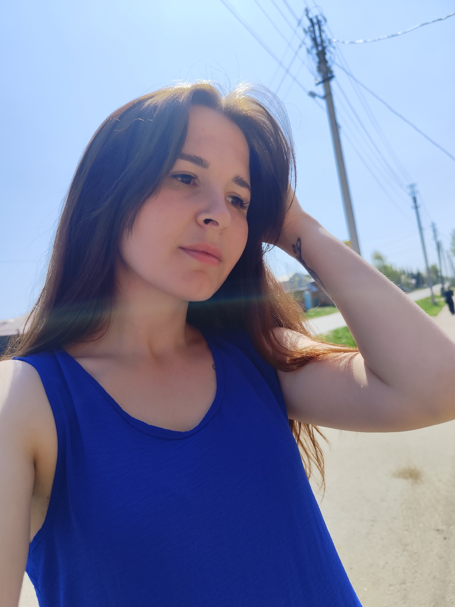 A woman with long brown hair, wearing a blue tank top, is standing on a street in what appears to be a rural or suburban area. She has her right hand raised to her head, suggesting she might be adjusting her hair. The background features utility poles with wires, trees, and a road. The photograph is taken during the daytime under bright sunlight. The location is Фоки, Russia, as indicated in the prompt.