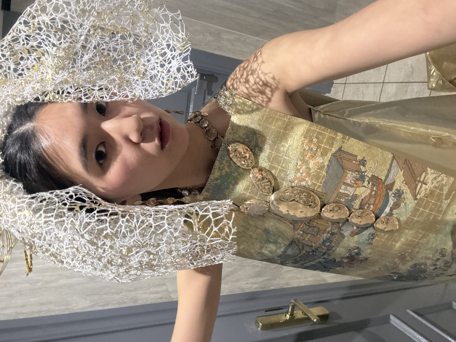 A young East Asian female is depicted indoors, in what is identified as Paris, France, posing for a self-portrait, holding the camera with her right hand. She is adorned with an elaborate ensemble. Her dark hair is pulled back, partially concealed by a prominent, structured headpiece made of white, web-like material interwoven with gold-toned fragments, casting a distinct net-like shadow on her right shoulder and arm. She wears long, dangling earrings composed of multiple clear, faceted beads, and a large, multi-component necklace featuring several irregular, hammered gold-toned metallic pieces arranged vertically down her torso. Her sleeveless garment consists of a shimmering gold fabric, overlaid on her left side with a panel depicting a traditional East Asian landscape or genre scene, including figures in boats and architectural structures, rendered in muted greens, blues, and browns over a subtle grid pattern. She looks directly at the camera with a neutral expression. The background includes a light-colored wall or door behind her left side, fitted with a brass-toned lever door handle, and a darker, less distinct area behind her right side. Illumination appears to be from an artificial light source, positioned to the front-right.