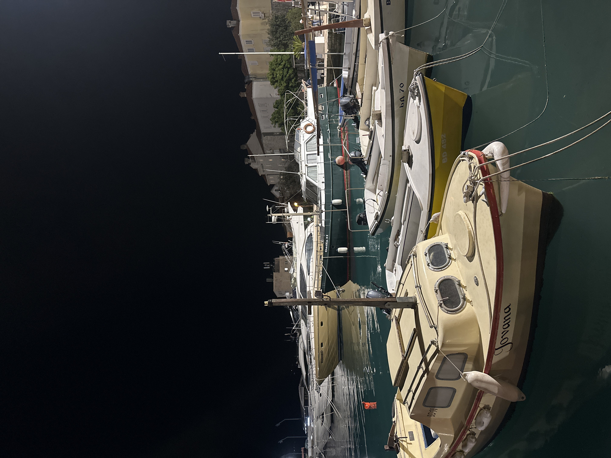 The image captures a serene night scene in a harbor in Budva, Montenegro. Numerous boats and yachts of varying sizes are docked along the waterfront, illuminated by artificial lights against a completely dark night sky.
In the foreground, on the right, is a cream-colored boat named "Jovana," written in cursive script on its hull, featuring a distinctive red trim. The registration "BD 114" is partially visible near its stern. Hanging fenders and multiple portholes are visible on its cabin. Adjacent to it are a white boat marked "BD 70" and a smaller yellow dinghy with the registration "BD 482."
Further into the harbor, a prominent dark green cabin cruiser is docked, with "Gathea II" visible on its stern. A man is discernible on the deck of this boat. Behind it, a larger, lighter-colored yacht named "ZORIS" is also moored. The calm water of the harbor reflects the lights from the boats and the surrounding area.
On the left side of the harbor, multi-story buildings with light-colored facades, characteristic of a Mediterranean town, line the shore. Green trees and foliage are visible between the structures. The overall atmosphere is peaceful, with the boats resting quietly under the night sky.