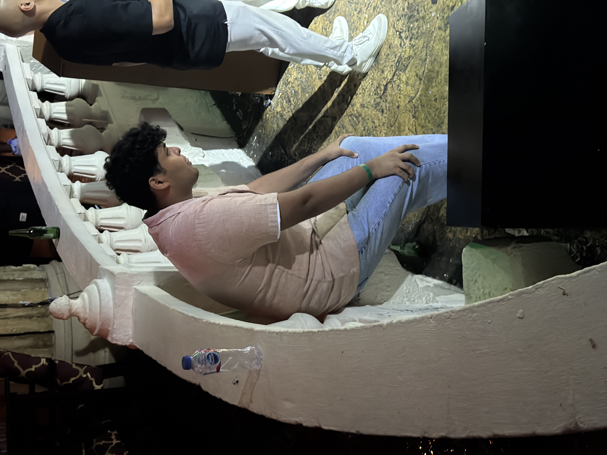 A young man wearing a light pink collared shirt and light blue jeans, with a green bracelet on his left wrist, is seated on a white curved ledge, looking downwards. To his right, a bald man in a black polo shirt and white pants stands, facing away from the camera. A plastic bottle of water is visible on the ledge to the left of the seated man. The scene appears to be outdoors at night, with artificial lighting casting shadows on a textured yellow surface. In the background, a white railing with decorative spindles lines the edge of a raised area. The context of Sharm El Sheikh, Egypt, is provided, suggesting a possible resort or entertainment venue setting.