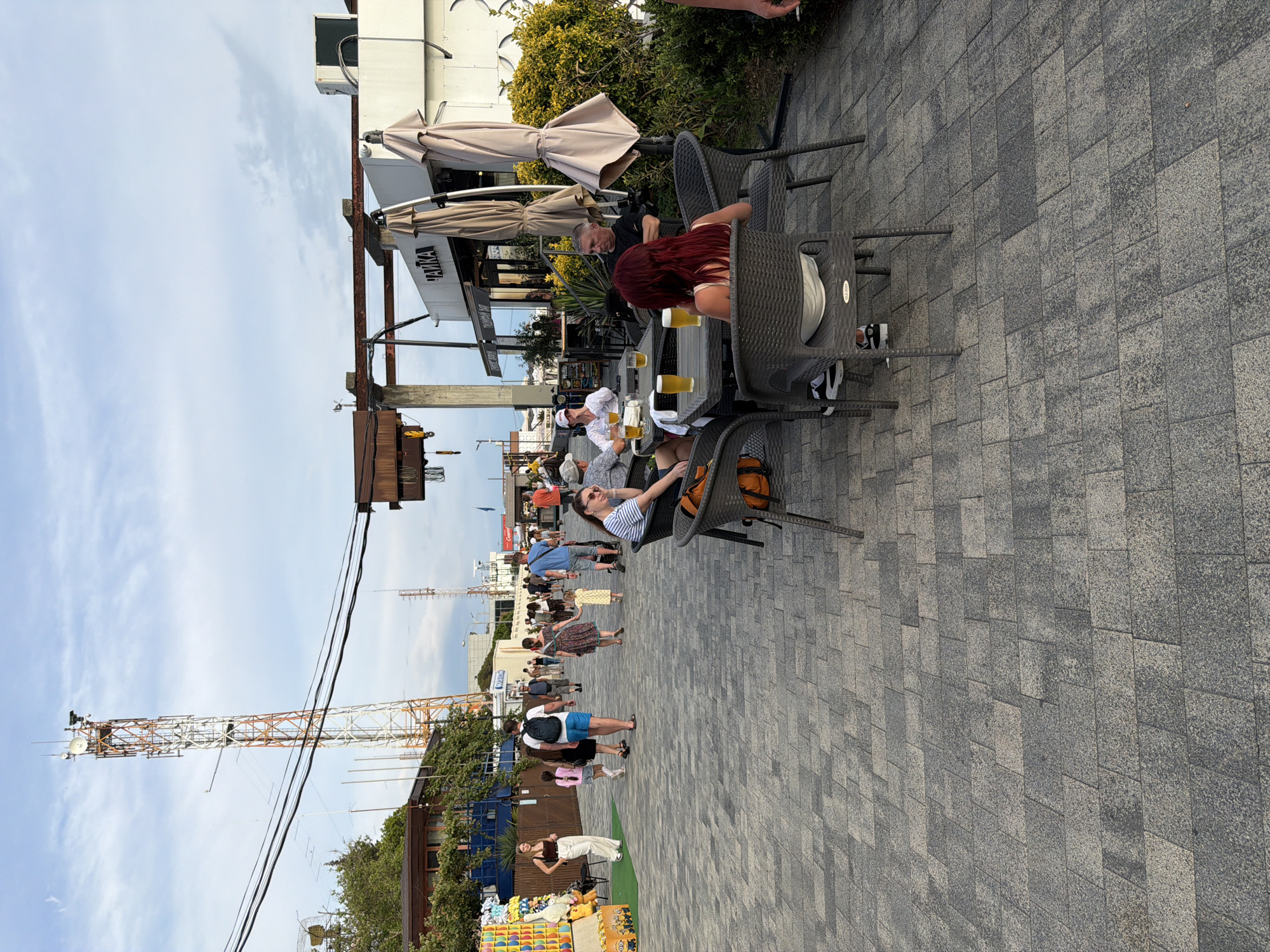 This is a vertical, wide-angle, eye-level shot of a bustling outdoor cafe and promenade area. The scene is populated with numerous people enjoying themselves. Several individuals are seated at tables, some with drinks, suggesting a leisurely atmosphere. Others are walking along the paved promenade, engaged in conversation or browsing.
In the foreground, several cafe tables and chairs are visible. One table has three glasses of beer and a woman with long, red hair is seated, facing away from the camera. Another table has a woman in a striped shirt sitting with her legs crossed, looking towards the right.
Further back, a street vendor is set up with a display of colorful goods, including sunglasses. The promenade is paved with grey rectangular stones. In the background, there is a tall communication tower, a building with awnings, and lush green foliage. The sky is partly cloudy, indicating it's likely daytime, perhaps late afternoon or early evening.
The overall mood appears to be relaxed and social, with people enjoying the outdoor setting. The location is identified as Sochi, Russia. There is some visible text on a building awning that reads "RITUAL", though it is partially obscured. The image captures a vibrant snapshot of daily life in a seaside resort town.
