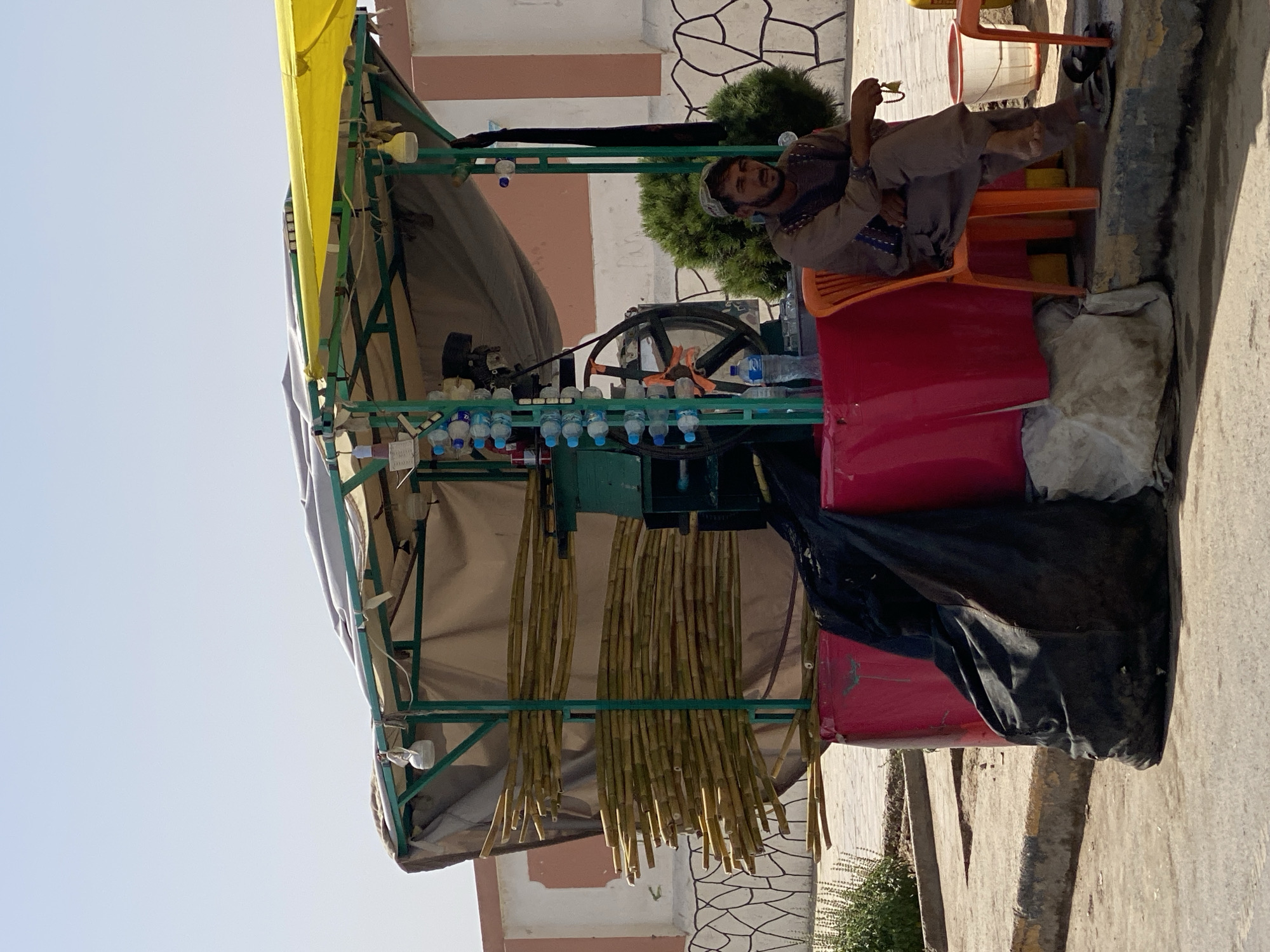 This image depicts a street-side sugarcane juice stand in Kandahar, Afghanistan. A man, appearing to be in his late 30s or early 40s, is seated on an orange plastic chair to the right of the stand. He is wearing a traditional shalwar kameez in muted colors and has a prayer bead rosary in his right hand. The stand itself is a makeshift structure with a green metal frame and a yellow canopy. Bunches of sugarcane stalks are prominently displayed on the left side. Behind the sugarcane, a sugarcane juicing machine is visible, along with a rack of plastic water bottles. A large red plastic container forms the base of the stand, with a black tarp draped over its side. The ground is paved with concrete and asphalt, with a curb separating the stand from the road. In the background, a low wall with a decorative pattern separates the street from a building. The sky is clear and bright, indicating a sunny day. The overall scene conveys a typical urban environment in Kandahar, likely during daylight hours.