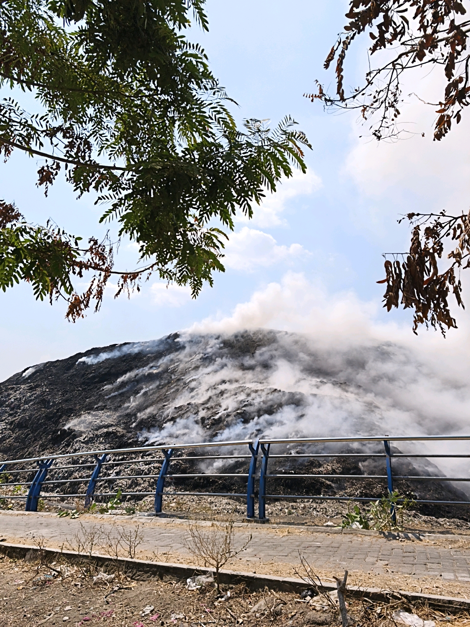 The image depicts a large pile of garbage, possibly a landfill, in Surakarta, Indonesia, with a significant amount of smoke rising from it, suggesting a fire or smoldering. In the foreground, a metal railing with blue supports separates a paved walkway from the garbage pile. The sky is partly cloudy, and trees with green and brown leaves are visible in the upper portion of the image. The ground in the foreground is strewn with litter and dry vegetation.