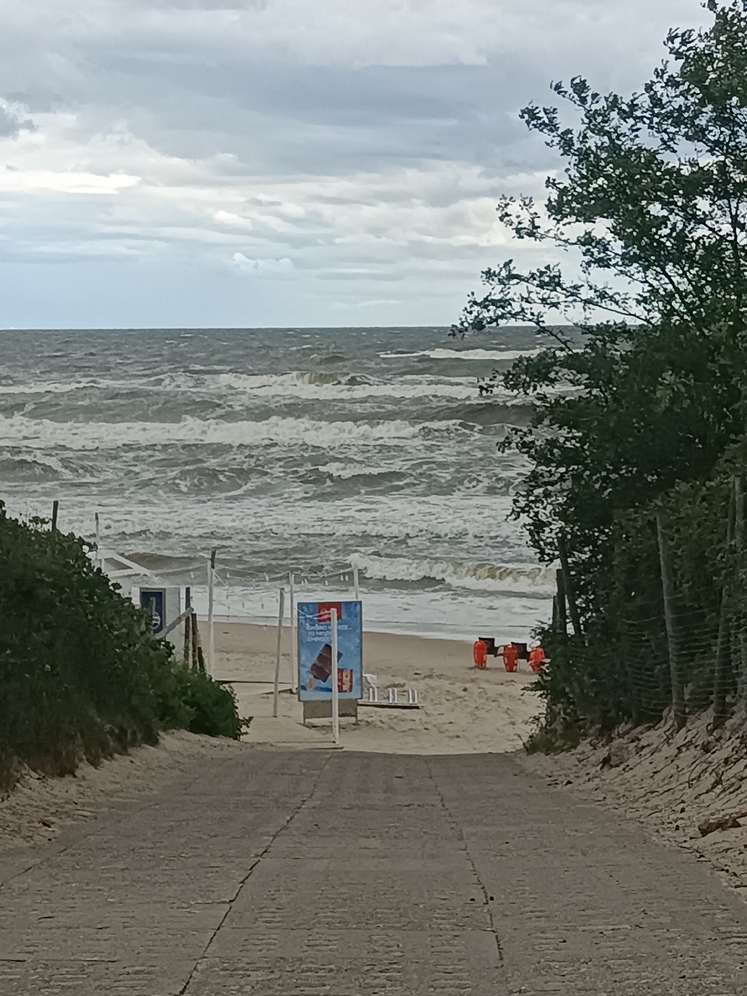 The image shows a grey, paved path leading downhill towards a sandy beach and a rough, choppy sea under a cloudy sky. On the beach, there are several orange life preservers arranged in a row, a small white beach hut with a blue door, and a blue sign displaying an image of an ice cream cone. Lush green foliage borders the path on both sides, with a dense tree on the right side of the frame extending towards the sky. The environment suggests a windy day at the Baltic Sea coast in Stegna, Poland, possibly during the off-season given the limited activity.