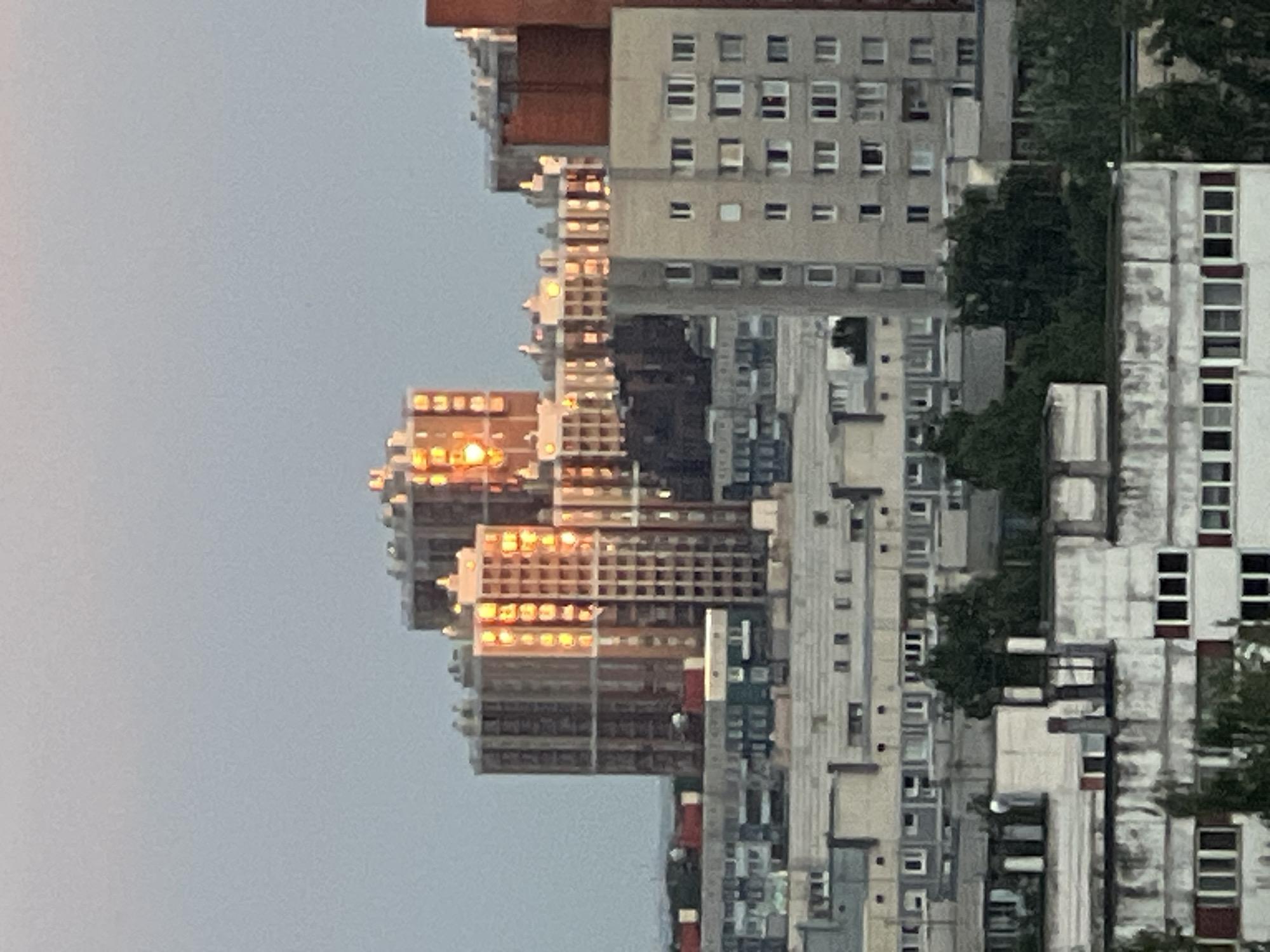 This is a rotated, medium-shot photograph of a dense urban area composed of apartment buildings. The buildings are constructed with concrete and glass, featuring numerous windows and balconies. Some of the buildings have a reddish-brown brick facade on the upper levels. Trees are visible interspersed between the buildings, suggesting an urban park or street greenery. The sunlight is reflecting off some of the windows, indicating it is either early morning or late afternoon. The sky is a pale blue, suggesting clear weather. There are no people or specific objects clearly identifiable within the scene, and no visible text. The overall impression is of a residential district within a city. The image appears to be taken from a slightly elevated perspective.