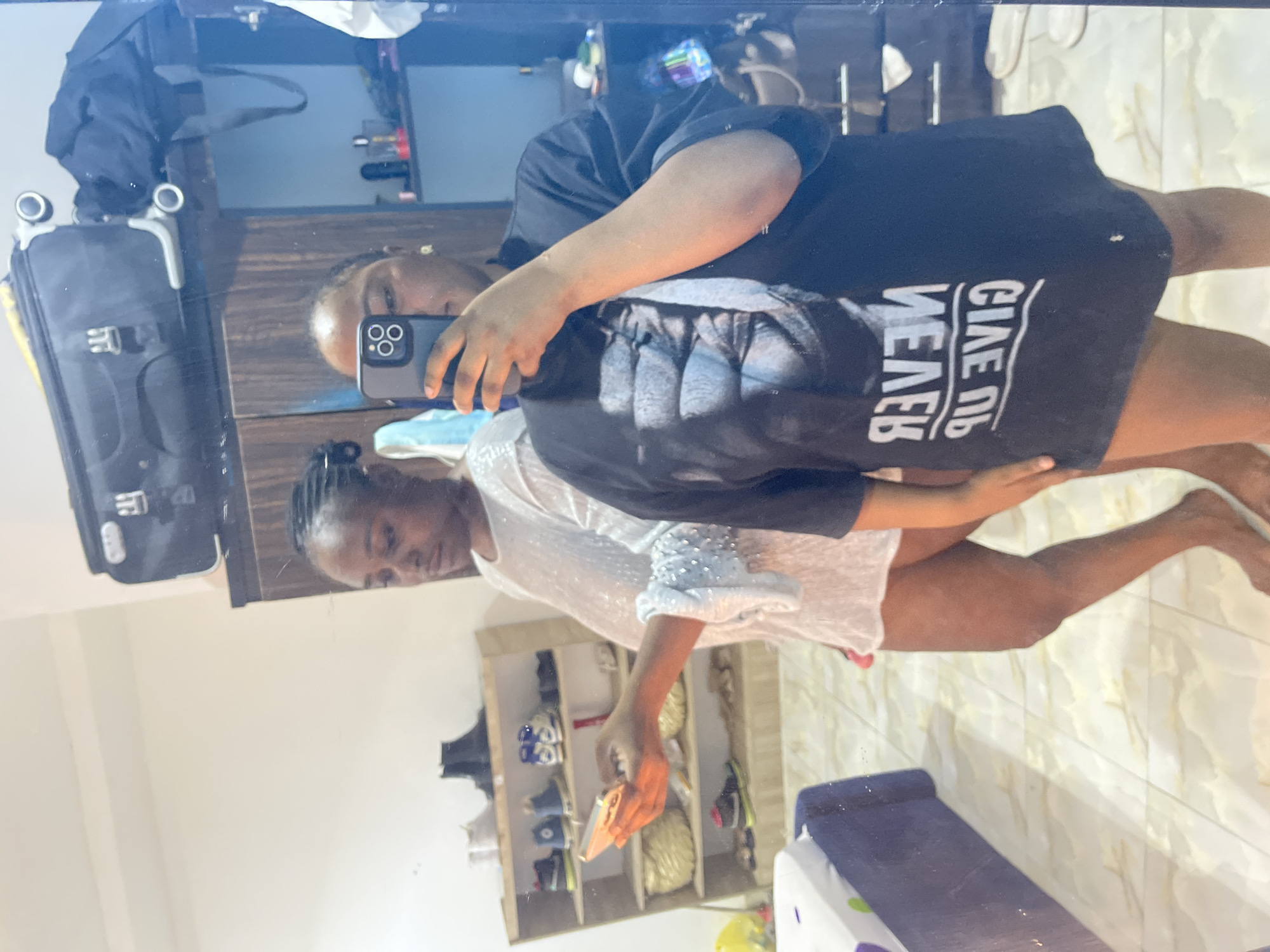 Two women are visible in a mirrored reflection. One is holding a phone, presumably taking the picture. She is wearing a black t-shirt with the reversed words "GIVE UP NEVER" written on it. The other woman is wearing a white, sequined top. There is a suitcase with wheels, a shoe rack filled with shoes, and a blue cushioned object visible in the background. The location is likely indoors, possibly a residence in Abuja, Nigeria, though there is no definitive way to ascertain location from the image.