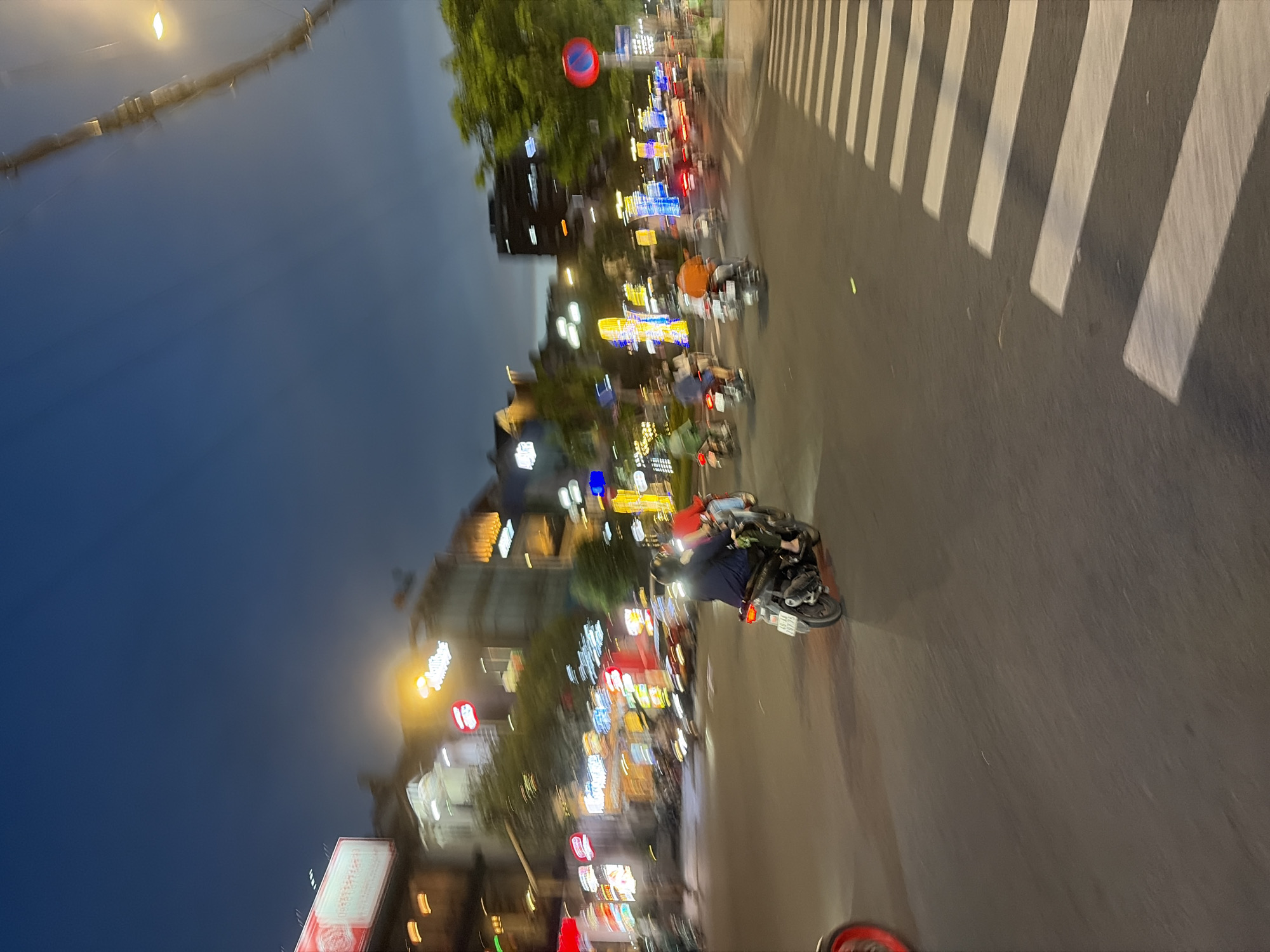 The image captures a dynamic, blur-filled street scene at night in Thủ Đức, Vietnam. The perspective is tilted, showing a dark blue sky above a bustling urban environment.
The main subjects are multiple motorbikes, some with riders, moving along a multi-lane road. In the foreground, a motorbike with a rider wearing a dark blue garment is visible, with its rear wheel and exhaust clearly defined despite the motion blur. Another rider in an orange top is visible further up the street. The heavy blurring of vehicles and lights suggests movement and activity.
The road features a prominent zebra crossing (crosswalk) on the right side, with its white stripes brightly illuminated. Buildings line both sides of the street, adorned with numerous, brightly colored neon signs and lights, which are largely blurred into streaks and glowing masses of color, indicating commercial establishments or eateries. Above the street, a red circular sign with a white horizontal bar, typically signifying "no entry" or "do not enter," is visible. Streetlights contribute to the overall illumination.
The scene depicts a typical evening or nighttime street in a Vietnamese city, characterized by a lively atmosphere created by the moving traffic and the vibrant glow of commercial signage. While specific text on most signs is illegible due to blur, their presence is a key visual element. The time of day is clearly night, with the sky transitioning from deep dusk to dark.