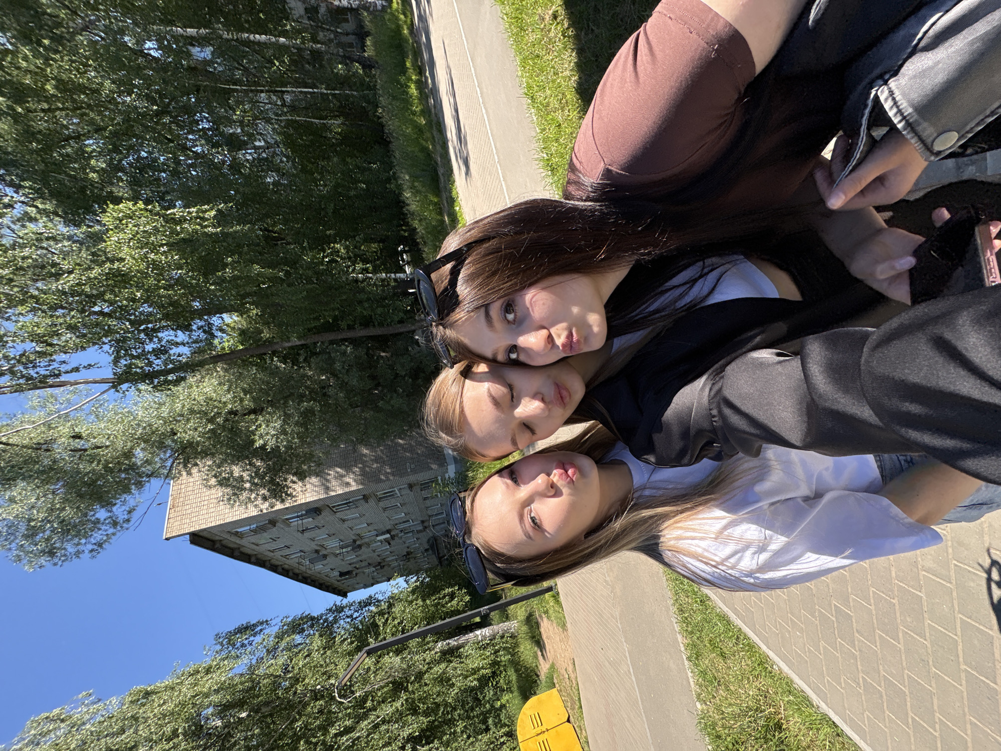 Three young women are posing for a selfie in an urban outdoor environment. The woman on the left has blonde hair and is wearing a white t-shirt. The woman in the middle has blonde hair and is wearing a black jacket over a white top. The woman on the right has dark brown hair and is wearing a brown t-shirt. All three women are puckering their lips for the photo. They are standing on a paved walkway next to a grassy area. In the background, there is a multi-story brick apartment building and trees. A yellow utility box is visible on the left side of the image. The sky is clear and blue. This scene is consistent with the residential areas found in the city of Chaikovsky, Russia, characterized by apartment blocks and green spaces.