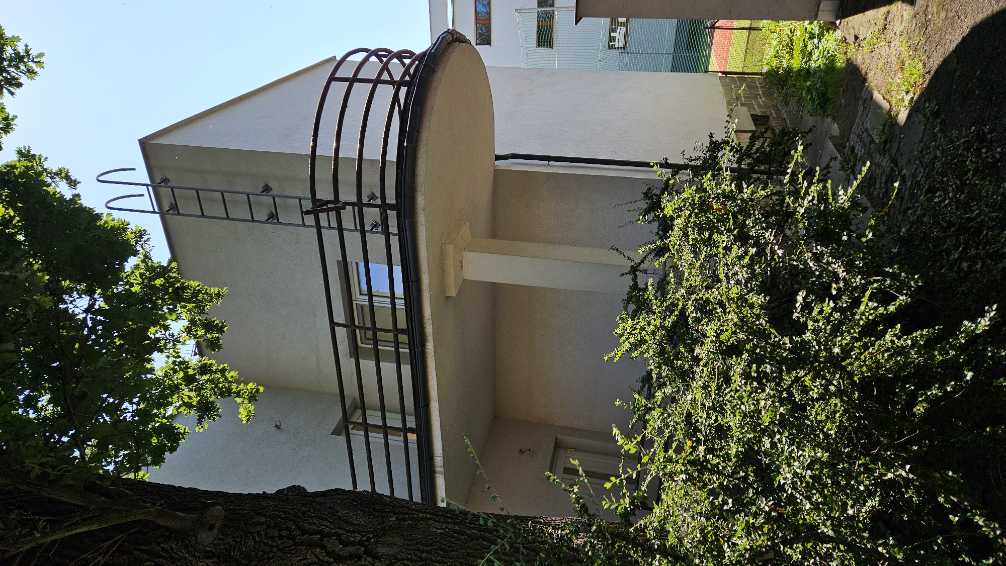 The image shows the exterior of a light-colored, modern building with a curved balcony. A metal ladder is attached to the side of the building, extending to the roof. Lush green bushes are in the foreground, partially obscuring a set of concrete steps leading down to a path. A large tree trunk is visible on the left side of the image. In the background, a tennis court with red and green surfaces is visible, enclosed by a fence. The sky is clear and blue, indicating a sunny day. The architecture and context suggest this is likely a residential or institutional building. This scene is located in Prešov, Slovakia, specifically at the former elementary school on Sibírska Street, which is now part of the secondary school complex.