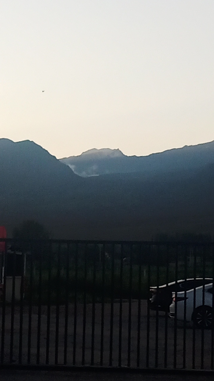 The image shows a wide view of a mountainous landscape at dusk or dawn. The sky is a pale, washed-out color, with a faint light suggesting the sun is low on the horizon. In the distance, dark, silhouetted mountains rise against the sky. A wisp of smoke or cloud is visible near the peak of one of the mountains, hinting at a possible natural phenomenon or human activity. In the foreground, a black metal fence runs horizontally across the frame, obscuring the lower portion of the scene. Through the fence, a dark, indistinct area suggests vegetation or perhaps buildings. On the right side, partially visible behind the fence, is the rear of a white car, its shape suggesting a modern hatchback or sedan. A small, dark object, possibly an airplane, is visible in the upper left quadrant of the sky. The scene appears to be located in a rural or semi-rural area, with the mountains serving as a prominent natural backdrop. Given the prompt, this scene is to be located in Choktal, Kyrgyzstan.