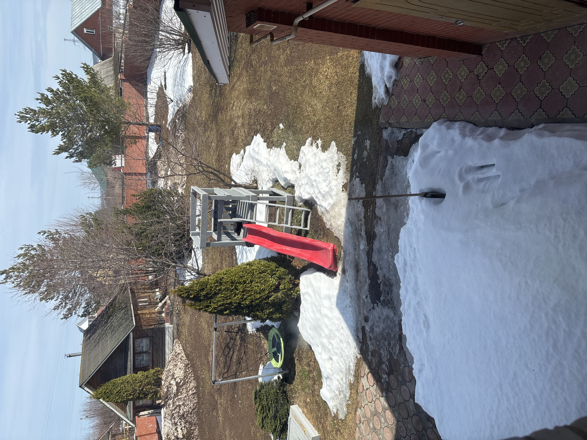 The image depicts a residential backyard in Ферма, Russia, during early spring. Patches of melting snow cover the brown grass. A children's playground with a red slide is visible, along with a swing set. The backyard is bordered by a brick pathway and a house. Several houses and trees are visible in the background. The sky is a clear blue.