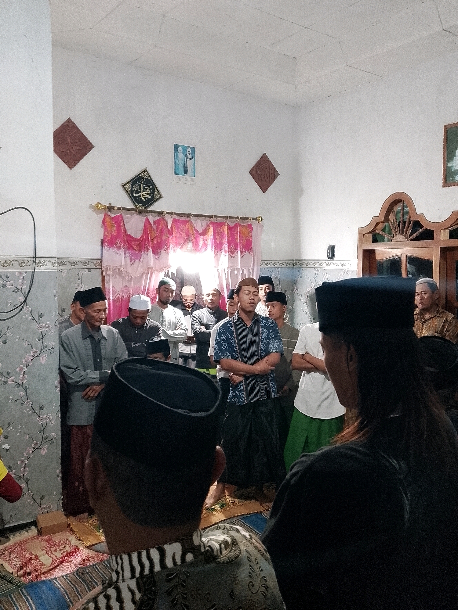 In the city of Rejoyoso, Indonesia, a group of men are gathered inside a room, seemingly for a religious ceremony. The room is adorned with traditional Islamic decorations, including calligraphy and framed artwork on the walls. A group of men, dressed in traditional Islamic attire such as peci hats and sarongs, stand or sit in prayer. Some are facing forward, while others are bowed in prayer. The room is lit by overhead lights, casting a warm glow on the scene. The overall atmosphere appears solemn and respectful, typical of a religious gathering.