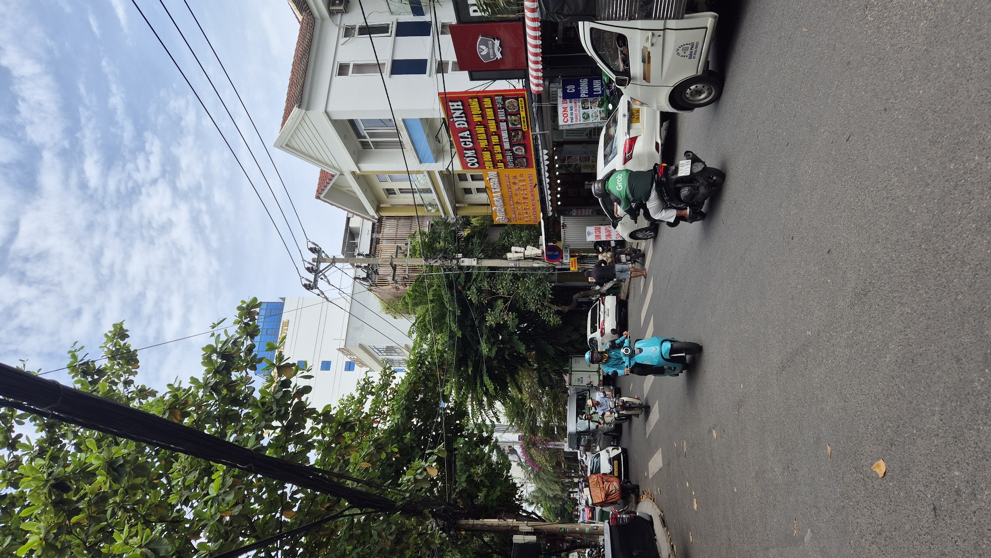 The image captures a street scene in the city of Đà Nẵng, Vietnam, on a cloudy day. A multi-lane asphalt road dominates the foreground, with pedestrians and various vehicles navigating the traffic. On the left, a lush green tree with visible leaves and branches overhangs the scene, partially obscuring electrical wires.
In the middle of the street, a person in a turquoise outfit rides a bright blue scooter, appearing to be moving towards the viewer. To the right of the scooter rider, another individual on a black motorcycle, wearing a green jacket with "Grab" branding, is also in motion. Further to the right, a white truck with a visible cargo bed is stopped, and on its side, text in Vietnamese reads "QUAN PHAT."
Several cars and motorcycles are visible further down the road. On the right side of the street, buildings line the road, some of which appear to be commercial establishments with prominent signage. One sign, in red and yellow, displays Vietnamese text, including "COM GIA DINH" (Family Meal).
The environment suggests an urban setting with a mix of residential and commercial properties. The sky is partially covered with white clouds, and the overall lighting indicates daytime. The presence of numerous motorcycles and scooters is characteristic of traffic in Vietnamese cities.