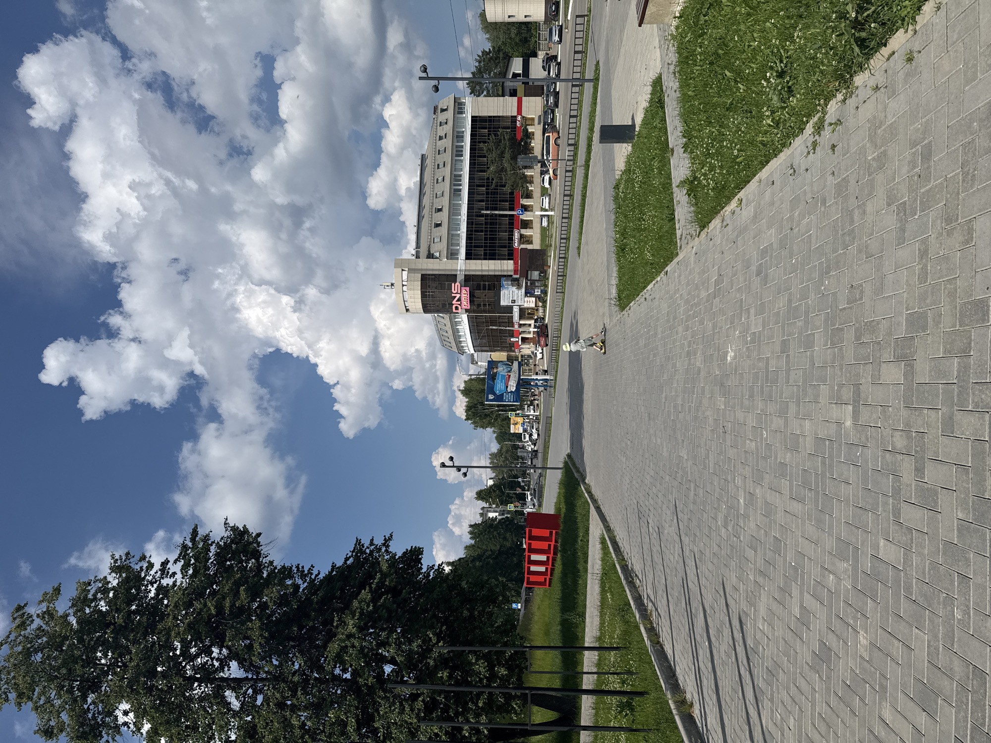 An outdoor, eye-level, medium shot captures a paved walkway in the city of Chaikovsky, Russia, with a modern, multi-story building in the background. The walkway, constructed of gray interlocking pavers, extends into the distance, bordered by a strip of green grass on the right. On the left, a large tree with dense foliage stands next to a series of black metal posts. Further back, a bright red modular structure with three white panels is visible.
Across the street, the modern building features a curved facade with extensive glass windows and a beige, angular upper section. A prominent neon pink sign reading "DNS Гайлер" is affixed to the building. Several cars are parked along the street in front of the building. In the mid-ground, a young child on a scooter rides along the sidewalk, facing away from the camera. The sky above is a vibrant blue, dotted with fluffy white cumulus clouds. The scene appears to be on a sunny day.