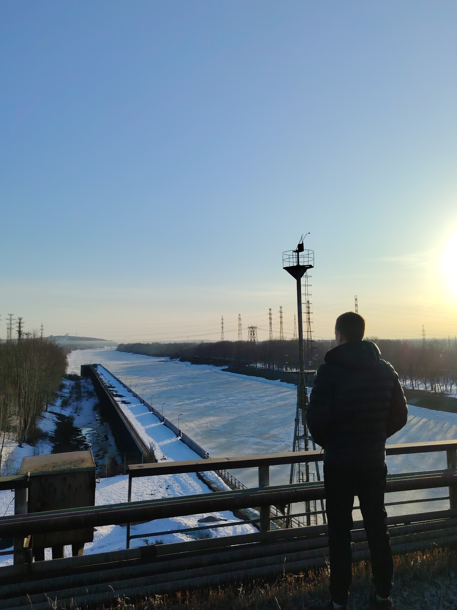 A man stands on a bridge in Chaikovsky, Russia, overlooking a partially frozen canal. He is wearing a dark jacket and trousers, facing away from the viewer, with his hands seemingly in his pockets. A black metal structure, possibly a navigational aid, stands near him. The canal is flanked by snow-covered banks and bare trees, with power lines visible in the distance. The sky is a clear gradient of blue and yellow, indicating either sunrise or sunset. There is snow on the ground in the foreground.