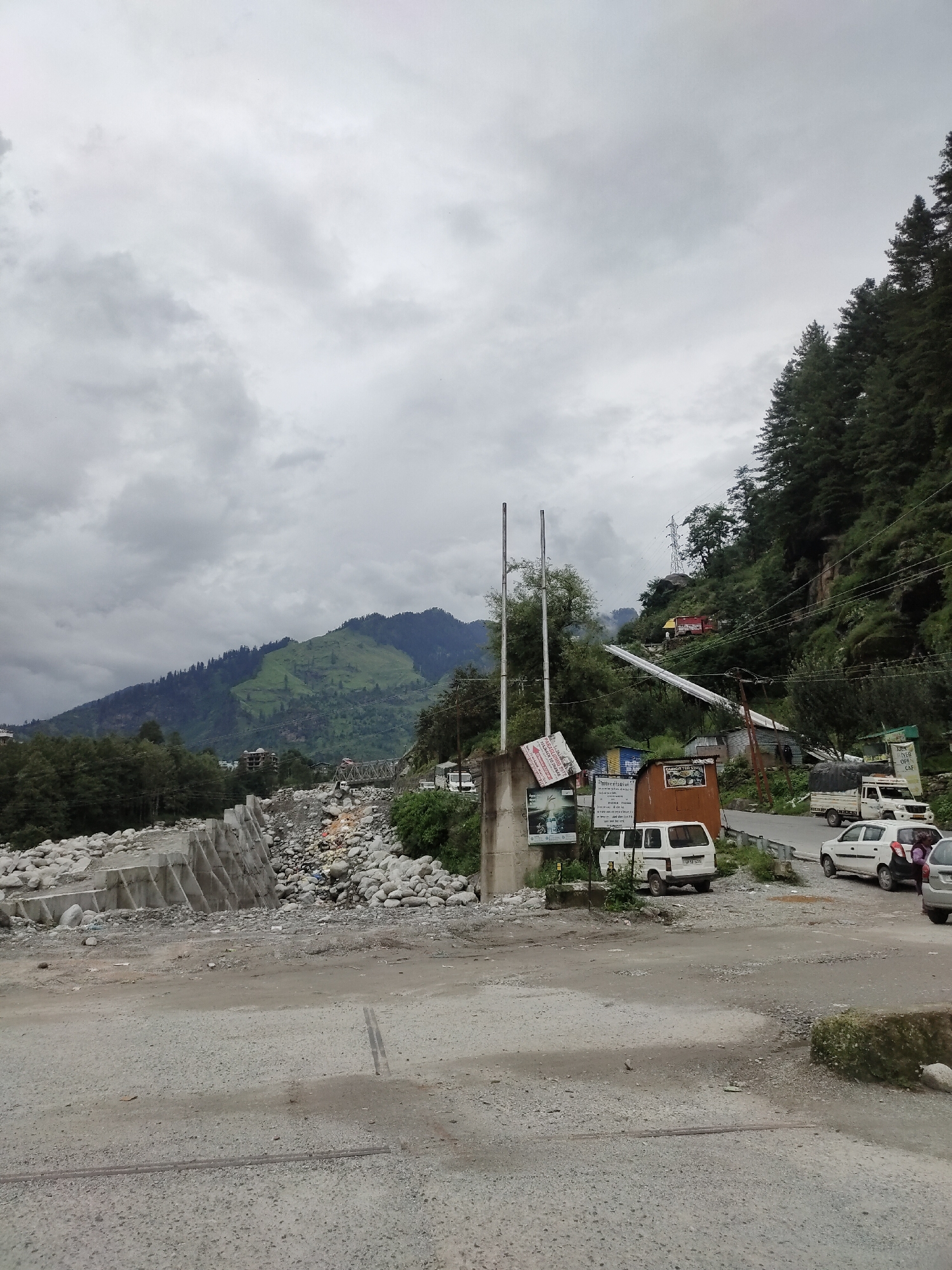 The image depicts a cloudy day scene in Barheta, India, with a focus on a road bordered by a rocky riverbed on the left and a densely forested hillside on the right. The riverbed features a concrete embankment with a diamond pattern texture, constructed with large rocks. Several vehicles, including white vans, a white car, and a truck with a tarped bed, are parked or driving on the asphalt road. Small structures, possibly shops or stalls, are visible along the roadside, some with signage. A bridge is visible in the distance, spanning across the valley. The hillside is covered with tall, dark green pine trees. The overall atmosphere is subdued, consistent with overcast weather.