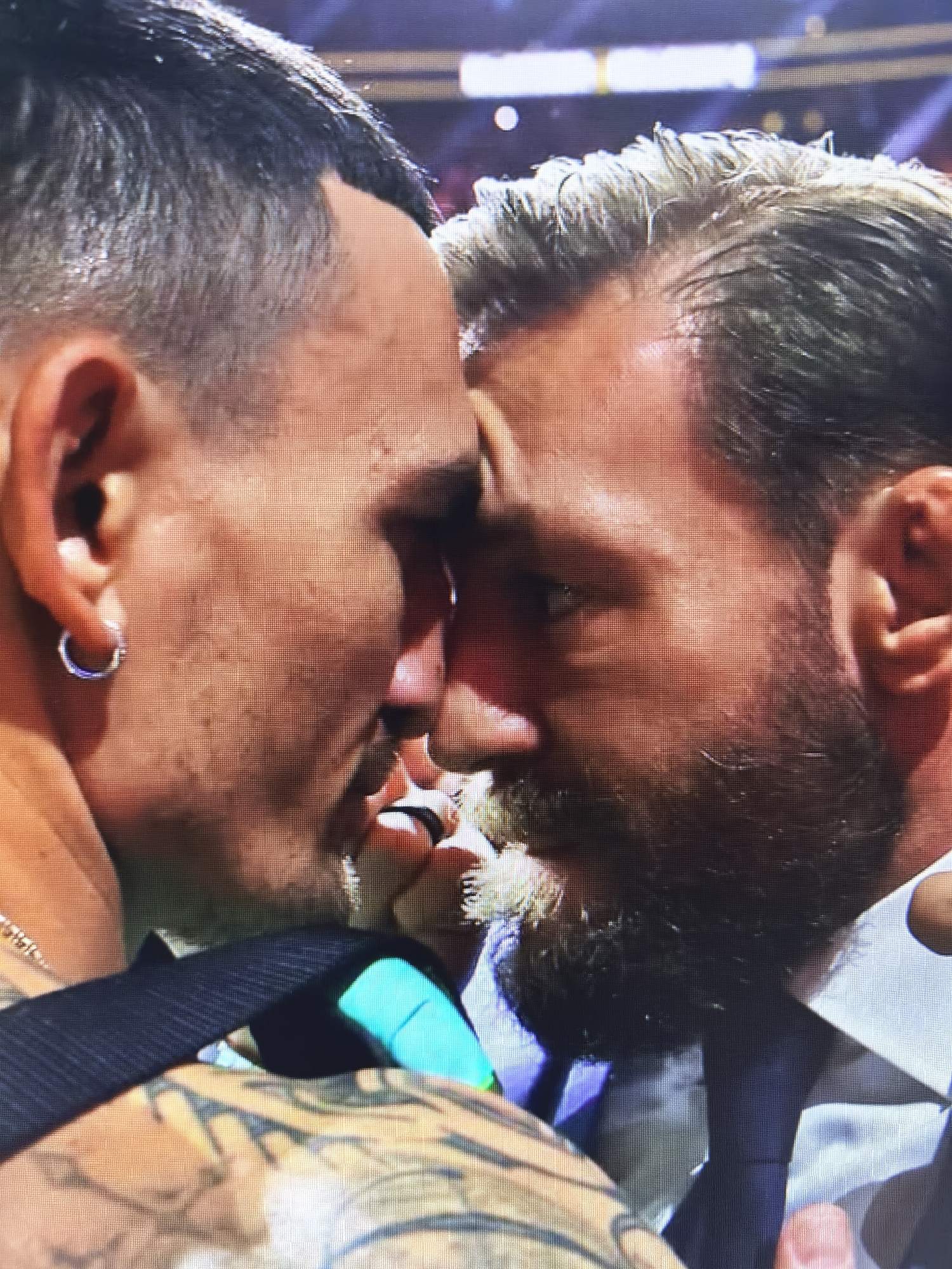 The image shows a close-up, head-to-head shot of two men. The man on the left has short, dark hair, an earring in his left ear, and visible tattoos on his arm and shoulder. The man on the right has lighter, possibly graying hair, a beard, and is wearing a white collared shirt. Their faces are very close, with foreheads and noses touching, suggesting an intense or confrontational moment. The background is blurred and appears to be an indoor setting with bright lights, possibly a stadium or event venue. There is no indication of the city of Fergana, Uzbekistan, in the image.