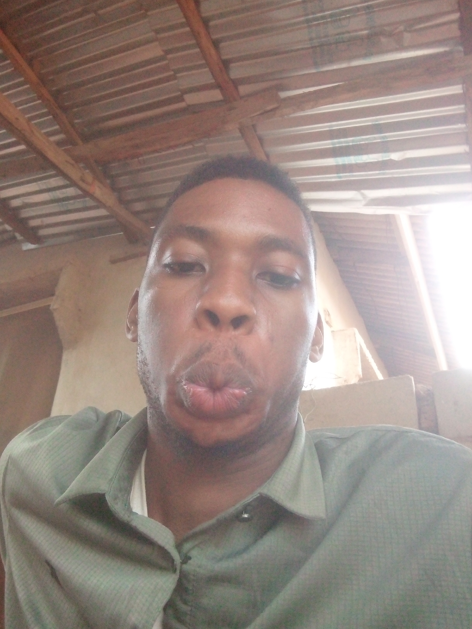 A man is taking a selfie, with his lips puckered slightly. He is wearing a button-down, collared shirt. Behind him is a corrugated metal roof with wooden supports. The location is Unknown, Unknown.