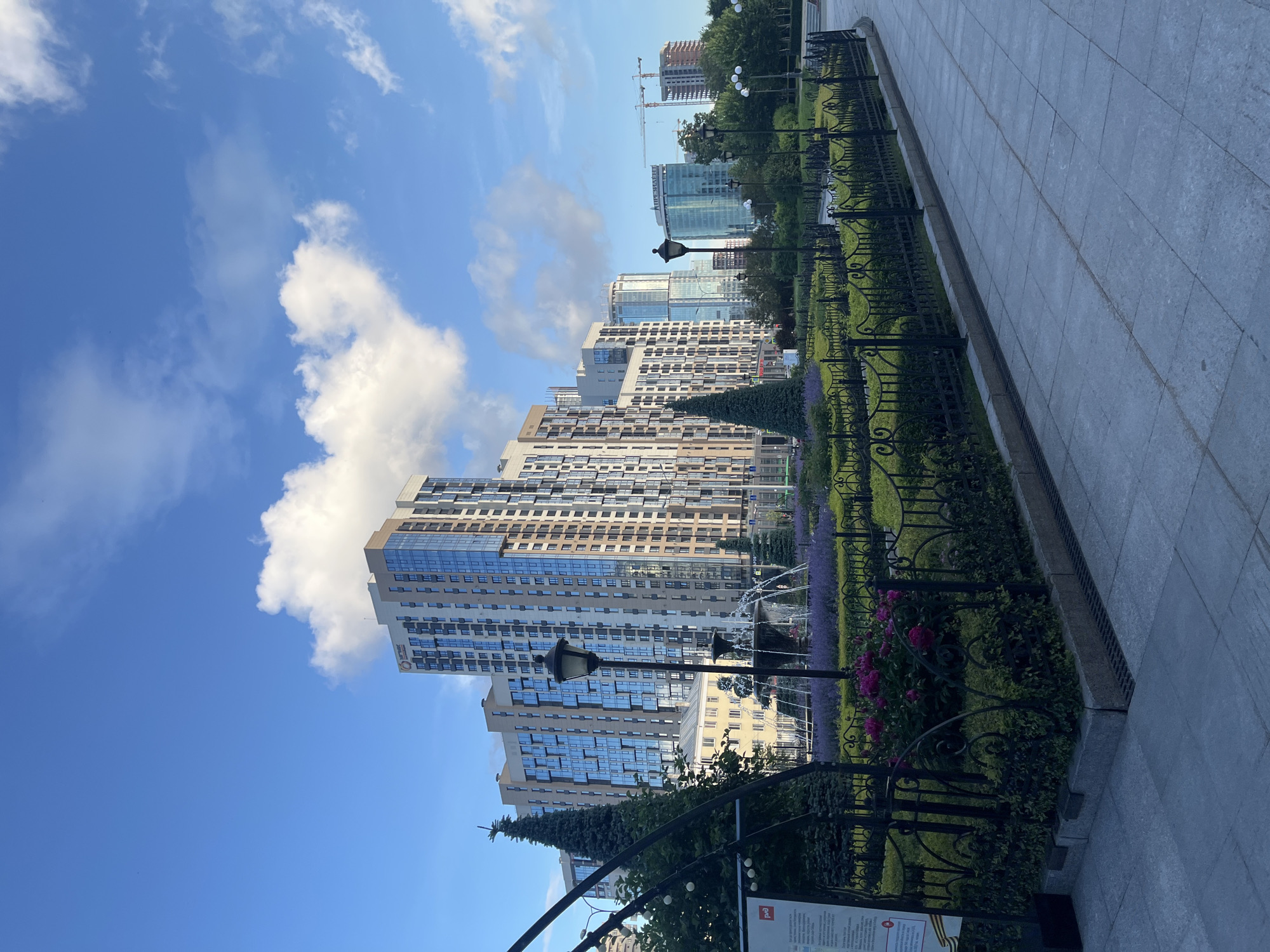 A wide shot captures a view of a city park with modern high-rise buildings in the background, under a bright blue sky dotted with white clouds. In the foreground, a paved walkway leads into the park, bordered by a manicured garden with a black wrought-iron fence. The garden features neatly trimmed green hedges, a row of purple flowers, and pink roses. A lamppost stands in the garden, and a fountain sprays water in the middle ground. Two conical evergreen trees flank the fountain. Beyond the garden, several tall residential and office buildings of varying architectural styles are visible, with some under construction, indicated by cranes. The overall scene depicts an urban landscape blending nature and modern development.