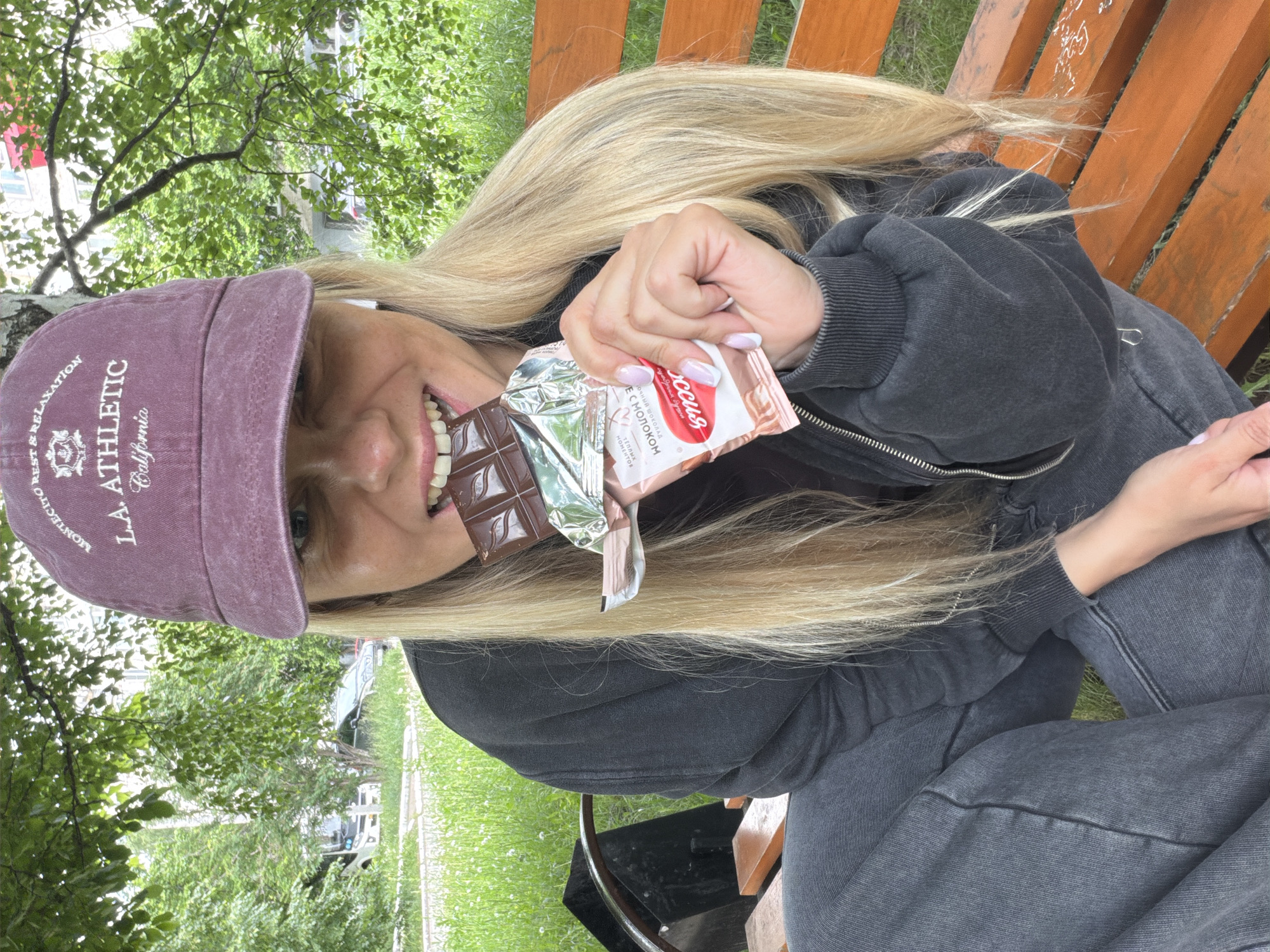 A young woman is depicted in a close-up, angled shot, sitting outdoors on a wooden slatted bench. She has long, blonde hair and is dressed in a dark, zippered hoodie and dark pants. On her head, she wears a purple baseball cap with white embroidered text reading "MONTECITO REST & RELAXATION L.A. ATHLETIC California."
The woman is smiling broadly, revealing her teeth, as she holds a partially unwrapped chocolate bar to her mouth, appearing to take a bite. The chocolate bar is dark brown, and its wrapper is white and pink with prominent red Cyrillic text. Visible words on the wrapper include "Россия" (Russia), "Щедрая Душа" (Generous Soul - likely the brand), "Молочный Шоколад" (Milk Chocolate), "С МОЛОКОМ" (With Milk), and "ТЕПЛЫХ МОМЕНТОВ" (Warm Moments). Her fingernails are manicured with a light pink base and white tips.
The background shows a vibrant outdoor scene with abundant green foliage from trees and grass. Some distant buildings are faintly visible beyond the trees. The natural lighting suggests it is daytime, possibly under an overcast sky. The overall impression is one of relaxation and enjoyment, with the woman happily snacking in a park-like setting in Votkinsk, Russia.
