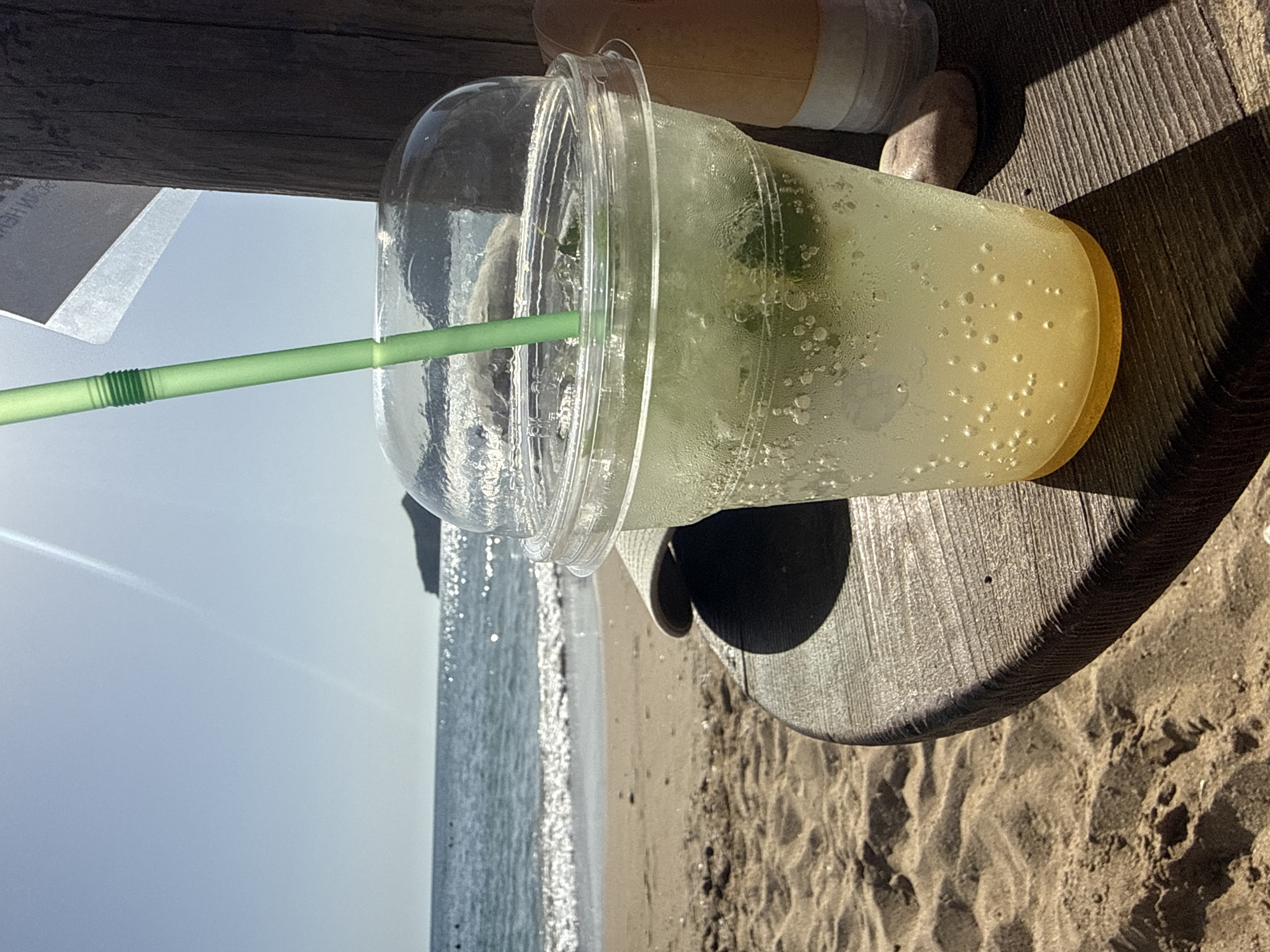 The image depicts a refreshing drink in a clear plastic cup with a lid and a green straw, placed on a wooden surface. The drink appears to be a light yellow-green color and contains ice cubes and mint leaves. The cup is beaded with condensation, suggesting it is cold. In the background, a sandy beach meets the sparkling blue sea under a bright, sunny sky. Waves are gently rolling onto the shore, and a distant island or landmass is visible on the horizon. The scene suggests a relaxed, tropical or coastal vacation setting. The location is identified as Agios Georgios, Greece. There are no people clearly visible in the image, though there are some dark shapes that could indicate distant figures.