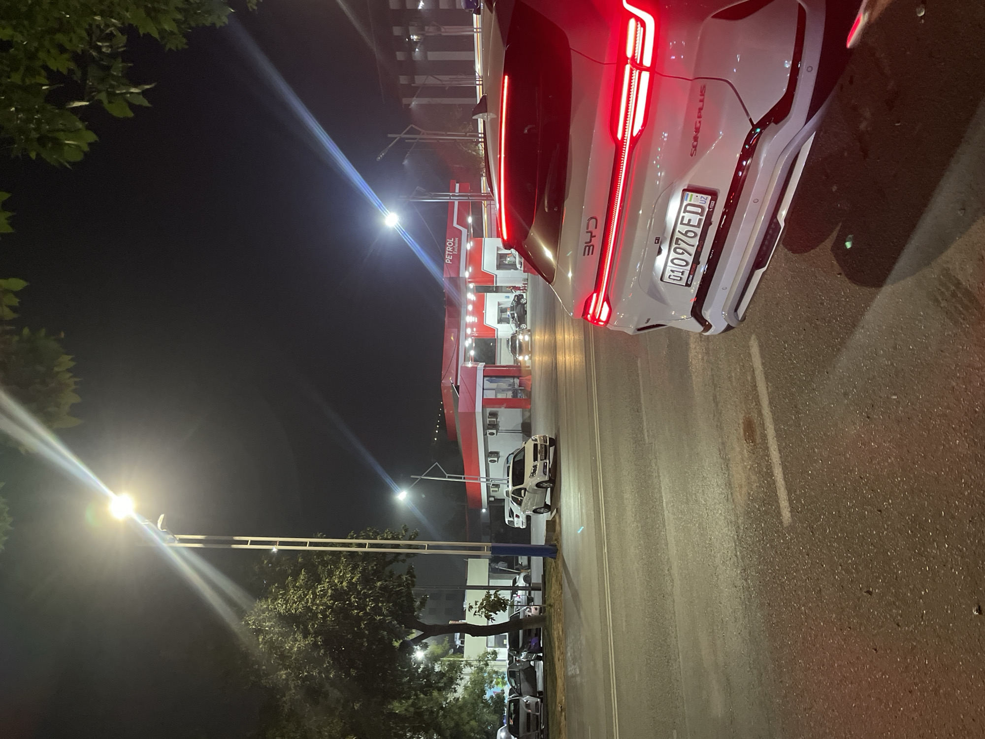 The image captures a nighttime scene at a Petrol gas station in Tashkent, Uzbekistan. In the foreground, the rear of a white BYD Song Plus electric vehicle is visible, its red taillights illuminated. The license plate reads "01 0976 ED" with the Uzbekistan flag emblem. The gas station, branded "Petrol Exclusive" with red and white architecture, is illuminated in the background, with several other vehicles parked and queuing. Streetlights cast a bright glow, creating lens flare effects. Trees with green foliage line the perimeter of the scene, and the asphalt road shows reflections of the lights. No people are clearly visible in the frame.