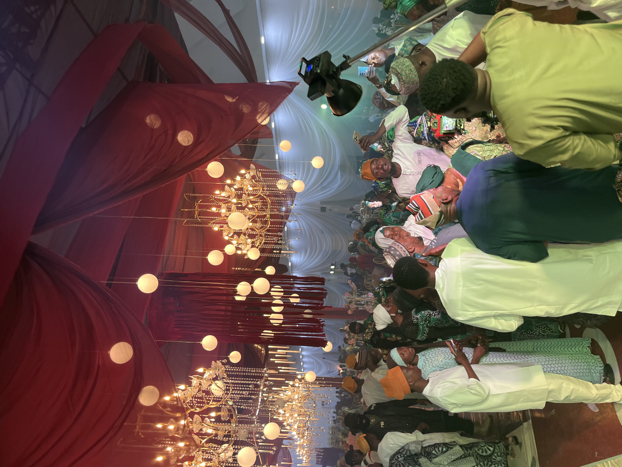 The image depicts a large gathering of people indoors, likely at an event such as a wedding or celebration. The environment is decorated with red draped fabric on the ceiling and elaborate crystal chandeliers, suggesting a formal or festive occasion. Several white spherical lights are also suspended from the ceiling. The attendees are dressed in traditional attire, predominantly in white, green, and patterned fabrics, with many men wearing caps or turbans. In the foreground, a cameraman is positioned with a professional camera on a stand, filming the event. Several individuals are interacting with the camera and the surrounding crowd, with some holding mobile phones to record. The overall atmosphere appears to be lively and celebratory.