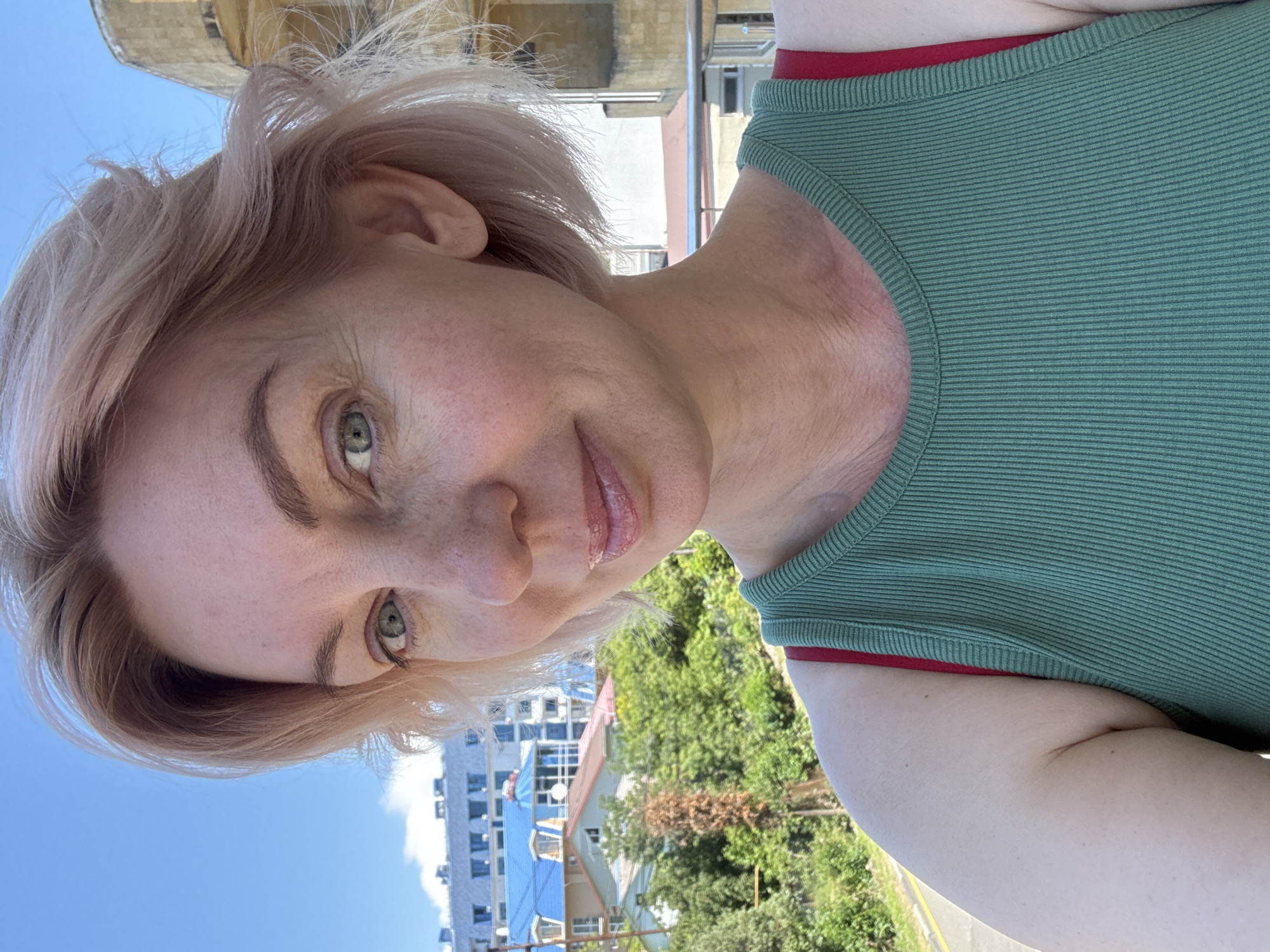A close-up selfie features a woman outdoors on a clear, sunny day in the city of Sirius, Russia. The woman, appearing to be in her late 30s to early 50s, has short, light blonde hair with a subtle pinkish tint, styled loosely with some strands appearing windblown. Her light blue-green eyes are directed towards the camera, and she exhibits a slight smile. Her fair skin shows visible freckles on her nose and cheeks, and some fine lines are present around her eyes and neck. Her lips appear glossy. She is wearing a green ribbed tank top, with a red bra strap visible over her left shoulder.
The background reveals a multi-layered urban and natural landscape under a bright blue sky with minimal cloud cover. In the distance, a large building with a prominent light blue roof and multiple windows is visible, characteristic of modern resort or residential architecture. Below it, several smaller buildings with various roof colors (red, brown, dark blue) are interspersed among dense green foliage, including trees and bushes, suggesting a hilly or vegetated terrain. To the right of the woman, a portion of a yellowish-tan building with a rounded upper structure and exposed concrete is partially visible. A paved road or path with a white line is discernible in the lower-left background, and a silver-colored railing is present in the lower-right, suggesting the woman is on an elevated platform or balcony.